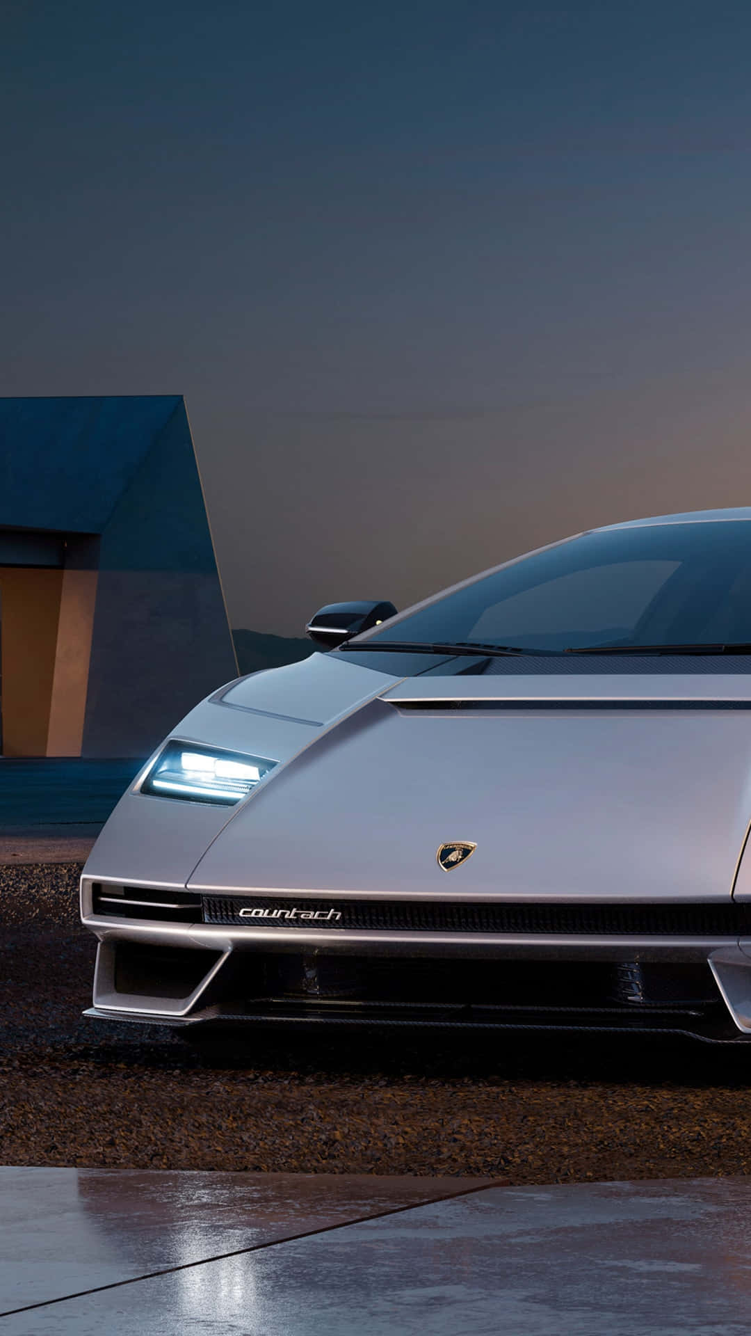 Pixel 3xl Lamborghini 桌布 壁紙 — 一輛時尚的銀色藍寶堅尼Countach停放在濕漉漉的路面上，在暮色中亮著車頭燈，背景是一座現代建築。