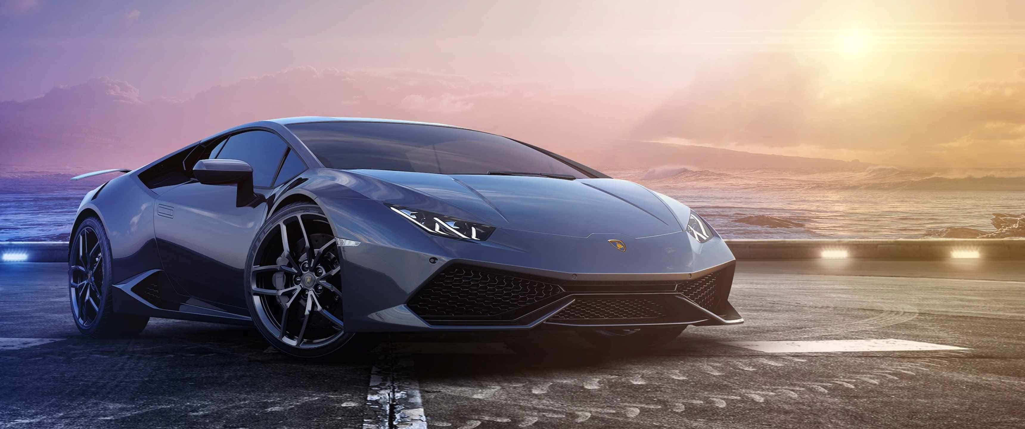 3440x1440p Lamborghini 桌布 壁紙 — 一輛時尚的銀色藍寶堅尼Huracán停放在日落時分的海岸公路上，車頭燈閃爍，天空戲劇性十足。