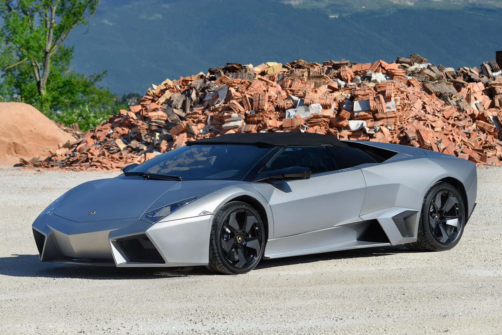 Lamborghini Reventón 桌布 壁紙 — 一輛時尚的銀色藍寶堅尼Reventon跑車停泊在碎石上，背景有一堆破碎的赤陶磚和綠樹。