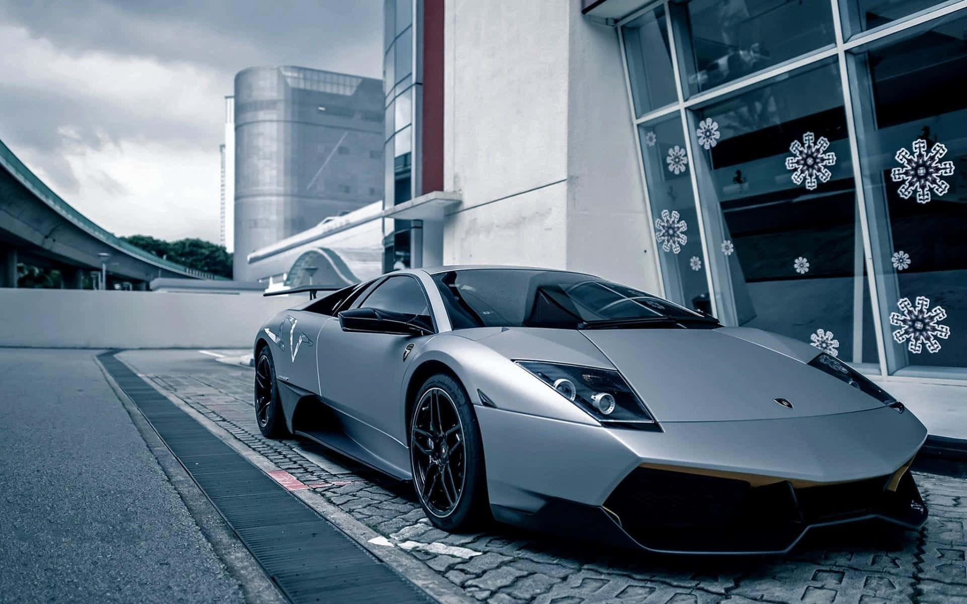 Lamborghini Murciélago 桌布 壁紙 — 一輛時尚的銀色藍寶堅尼Reventon停泊在鵝卵石路上，旁邊是一座現代建築，玻璃窗上有雪花貼紙，天空多雲。
