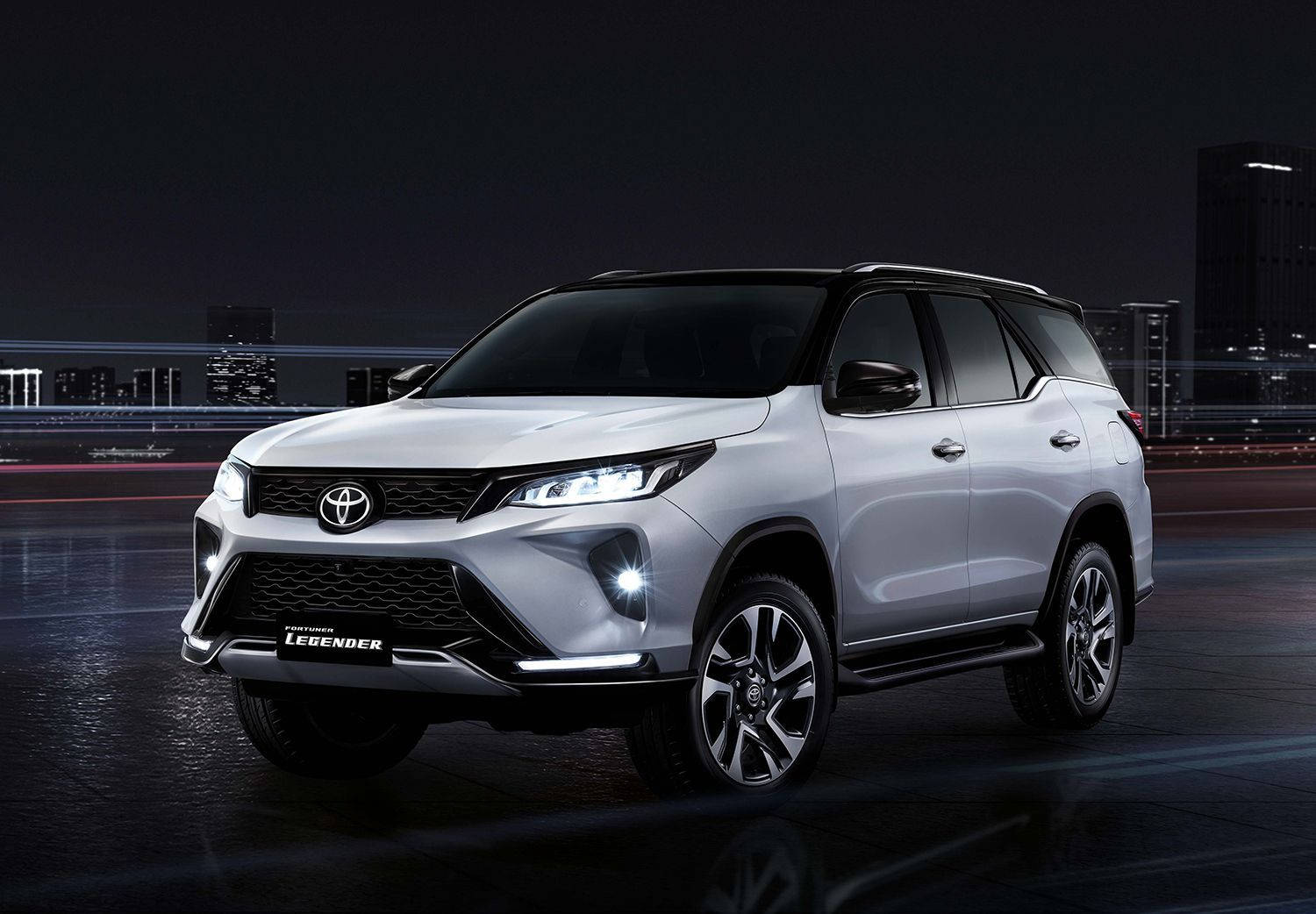 Toyota 桌布 壁紙 — 一輛時尚的銀色豐田 Land Cruiser Legend SUV 在夜間燈光下，於濕漉漉的城市道路上閃耀，背景是模糊的城市景觀。