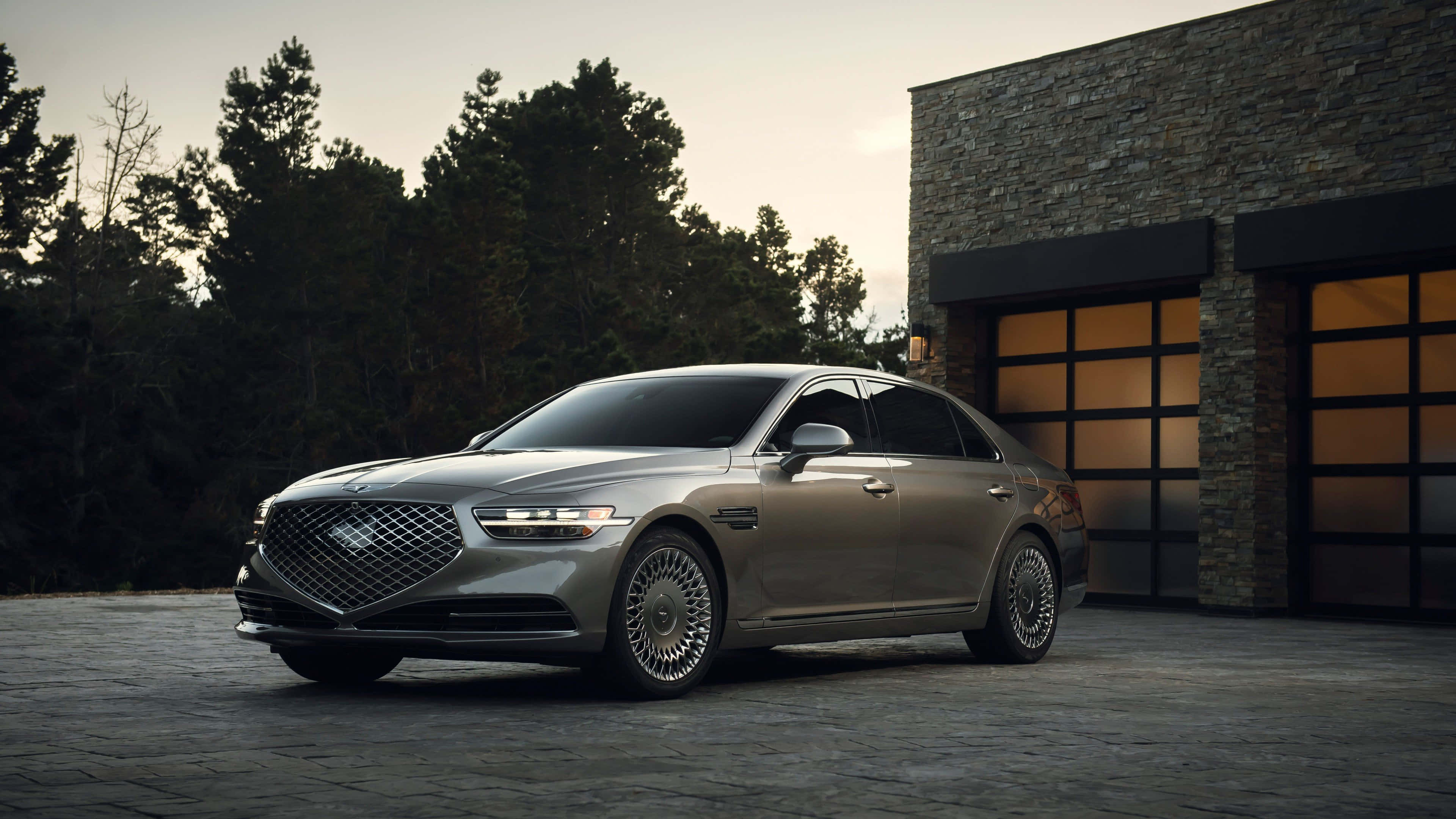 Genesis G90 桌布 壁紙 — 一輛時尚的銀色豪華轎車停泊在石頭車道上，旁邊是一座帶有發光車庫門的現代石頭建築。