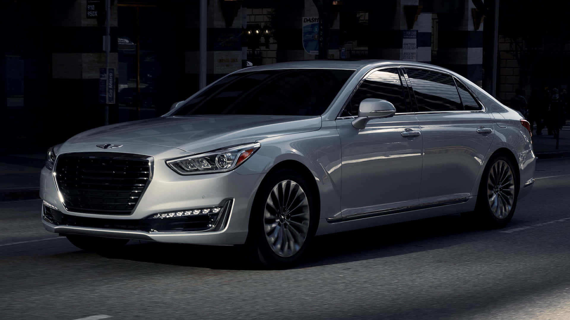 Genesis G90 桌布 壁紙 — 一輛時尚的銀色豪華轎車在夜間行駛於燈光昏暗的城市街道，具有反光表面和銳利的現代線條。