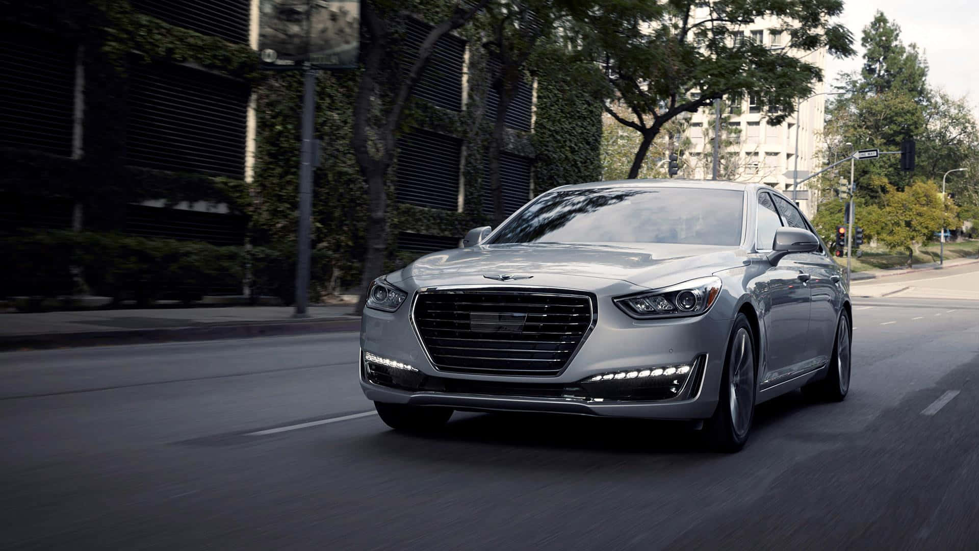 Genesis G90 桌布 壁紙 — 一輛時尚的銀色豪華轎車駛過綠樹成蔭的市區街道，背景是現代建築。