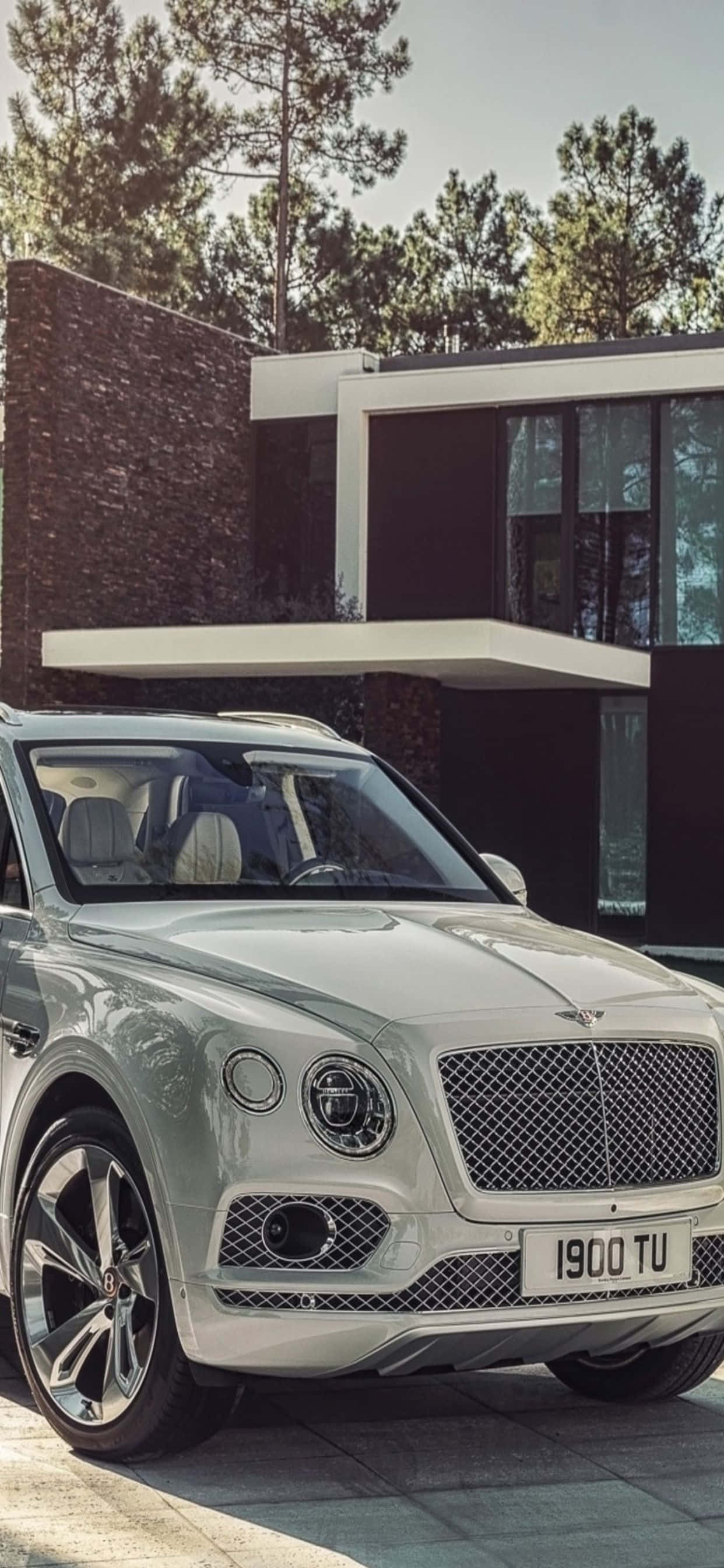 iPhone Xs Max Bentley 桌布 壁紙 — 一輛時尚的銀色賓利 Bentayga SUV 停在一棟現代石頭和玻璃房屋前，後面有高大的松樹。