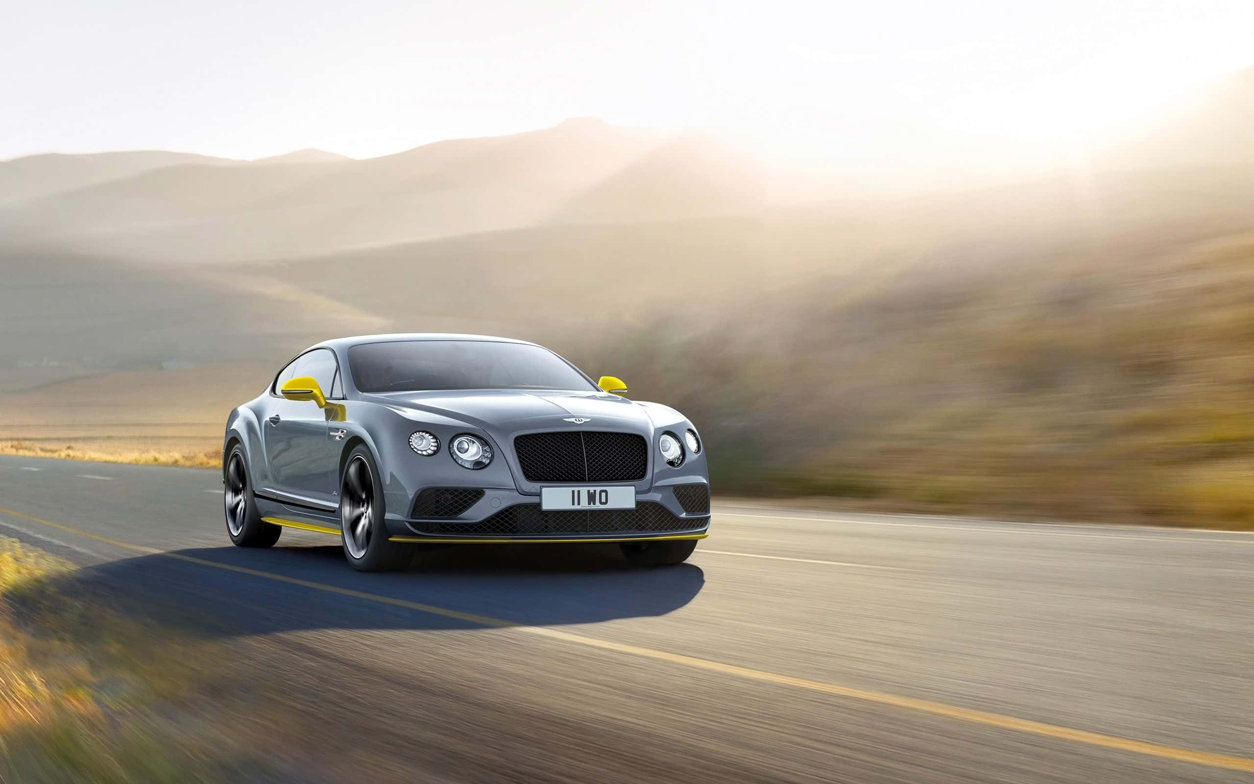 賓利 桌布 壁紙 — 一輛時尚的銀色賓利 Continental GT 在沙漠公路上疾馳，模糊的動態與日出時的金色山丘形成對比。