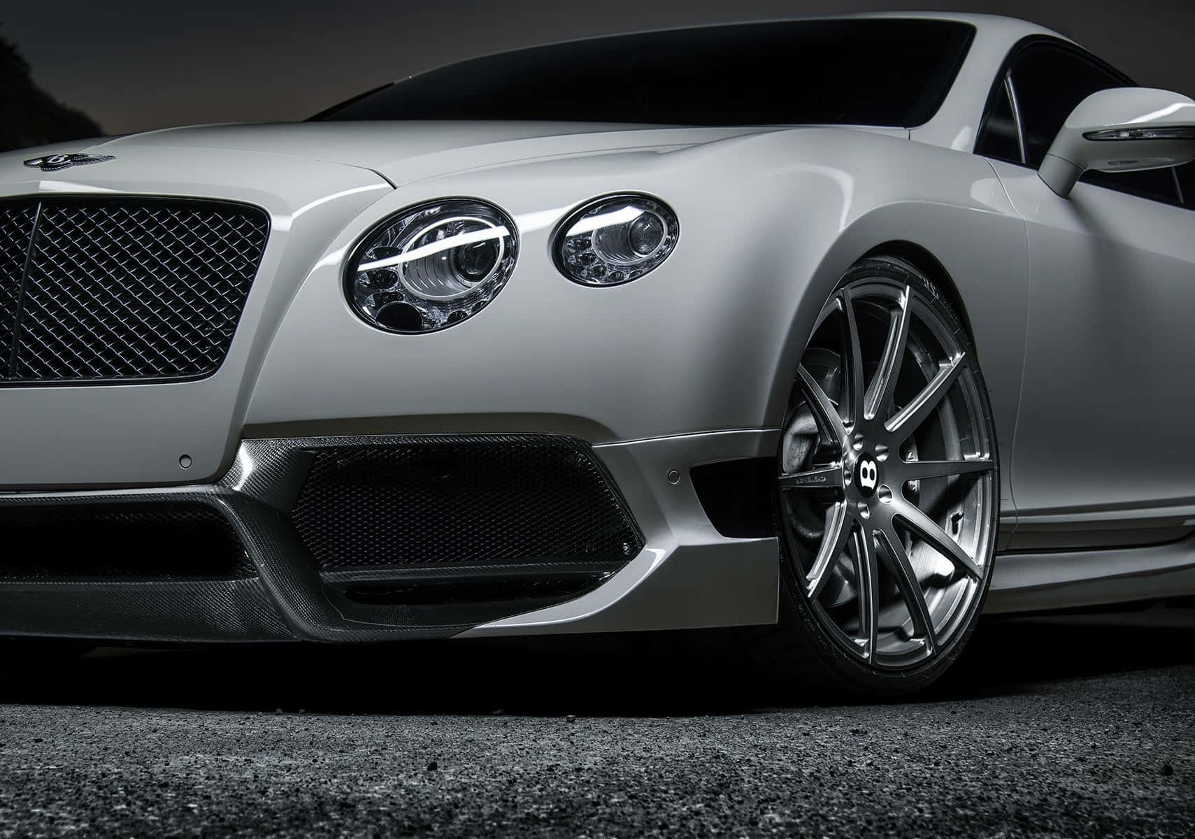 Best Bentley 桌布 壁紙 — 一輛時尚的銀色賓利 Continental GT 跑車，帶有碳纖維飾邊和發光頭燈，停在瀝青路面上，在戲劇性的燈光下。
