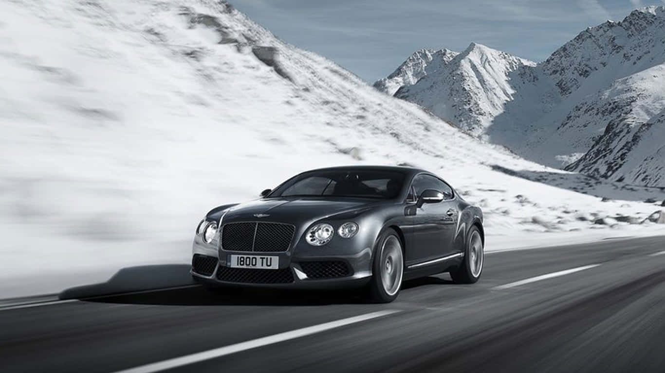 1366x768 Bentley 桌布 壁紙 — 一輛時尚的銀色賓利 Continental GT 在晴朗藍天下，沿著蜿蜒的山路疾馳，兩旁是白雪覆蓋的山峰。