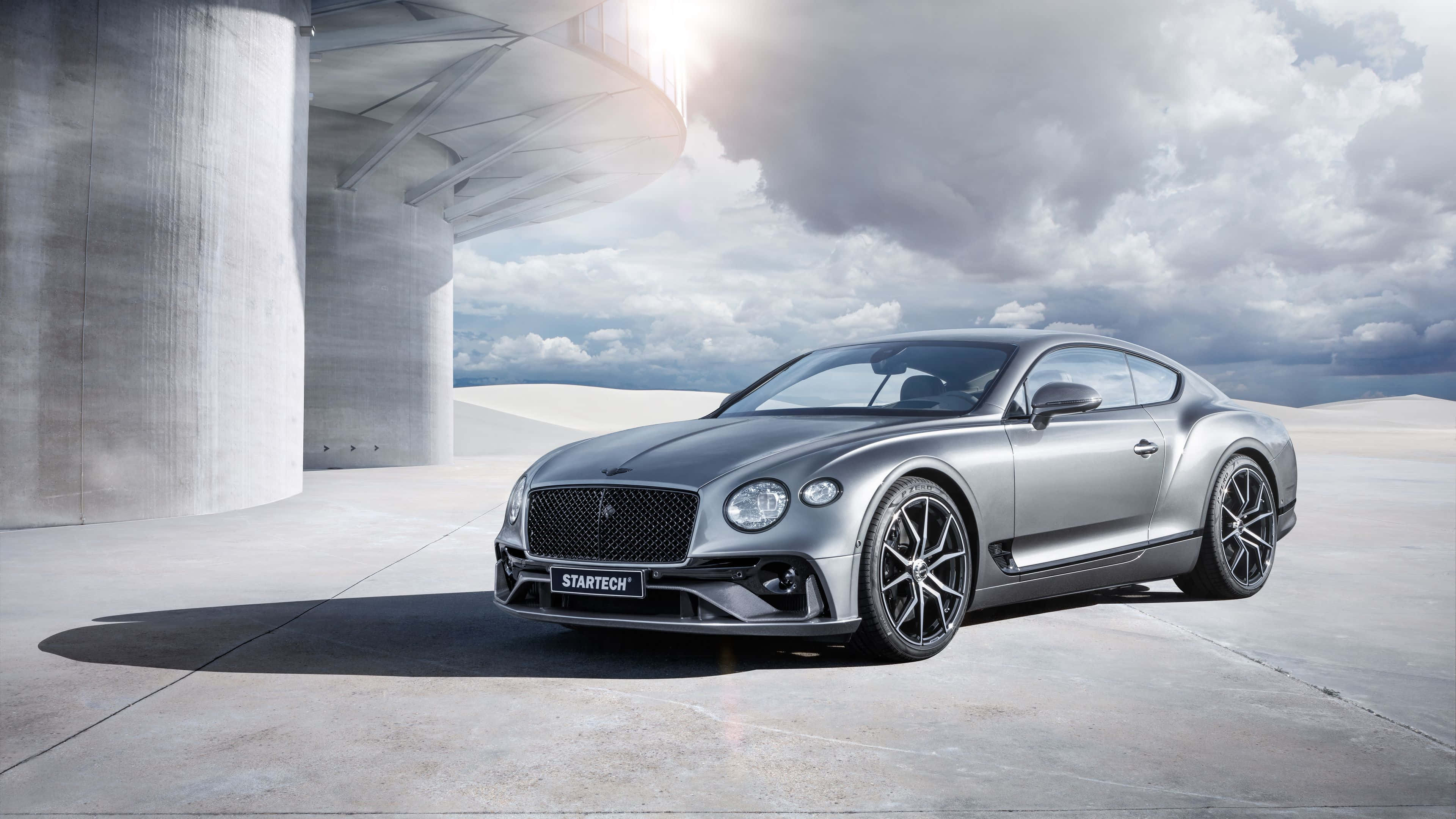 Best Bentley 桌布 壁紙 — 一輛時尚的銀色賓利 Continental GT 停放在現代混凝土結構下，襯托著戲劇性的多雲天空。