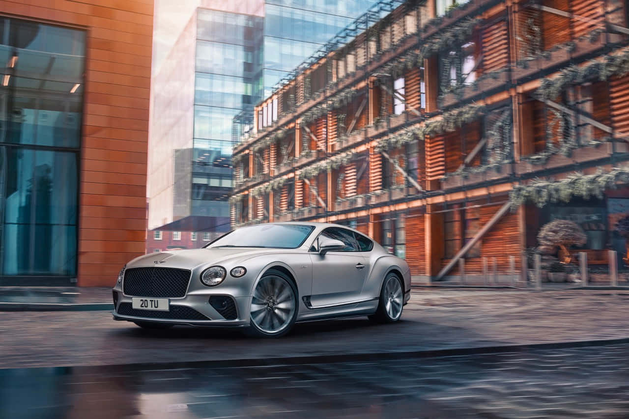 Bentley Mulsanne 桌布 壁紙 — 一輛時尚的銀色賓利 Continental GT 在柔和的日光下行駛在現代城市街道上，兩旁是玻璃和磚砌建築。