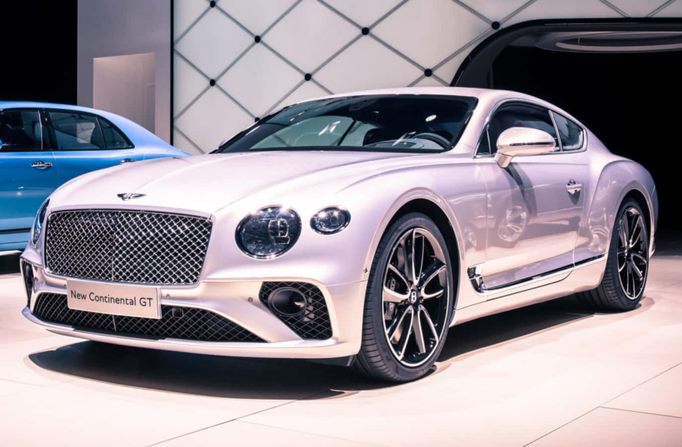 賓利 桌布 壁紙 — 一輛時尚的銀色賓利 Continental GT 在高級車展的明亮燈光下閃閃發光，背景有一輛藍色汽車。