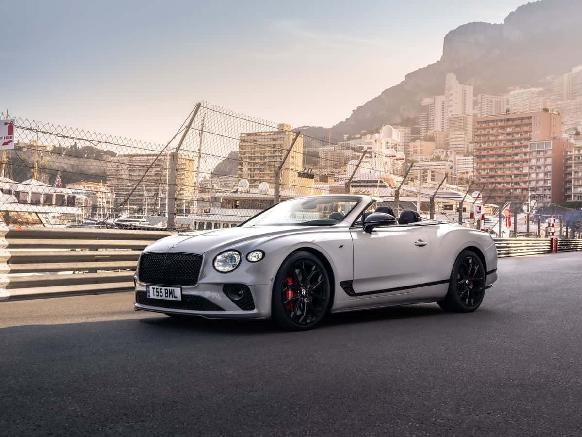 Bentley Continental Gt 桌布 壁紙 — 一輛時尚的銀色賓利敞篷車，帶有黑色飾邊和紅色制動卡鉗，停在俯瞰摩納哥海岸城市景觀的賽道上，正值黃金時刻。