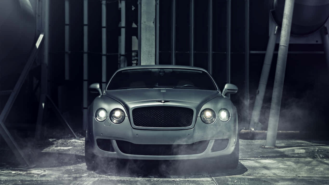 1366x768 Bentley 桌布 壁紙 — 一輛時尚的銀色賓利跑車在充滿煙霧的工業車庫中，車燈閃爍，鋼樑環繞。
