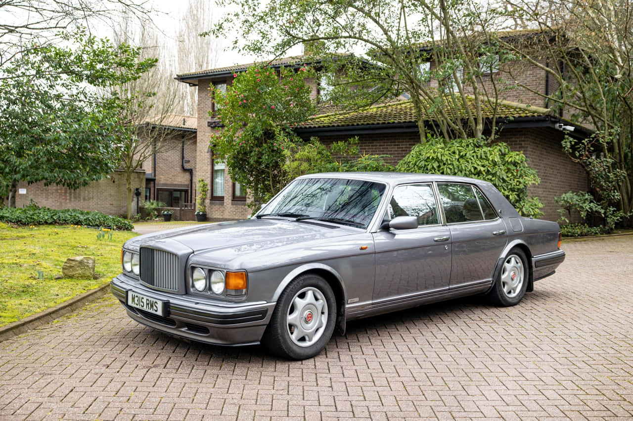 Bentley Turbo R 桌布 壁紙 — 一輛時尚的銀色賓利轎車停在磚砌車道上，車道前是一座被綠植環繞的磚房。