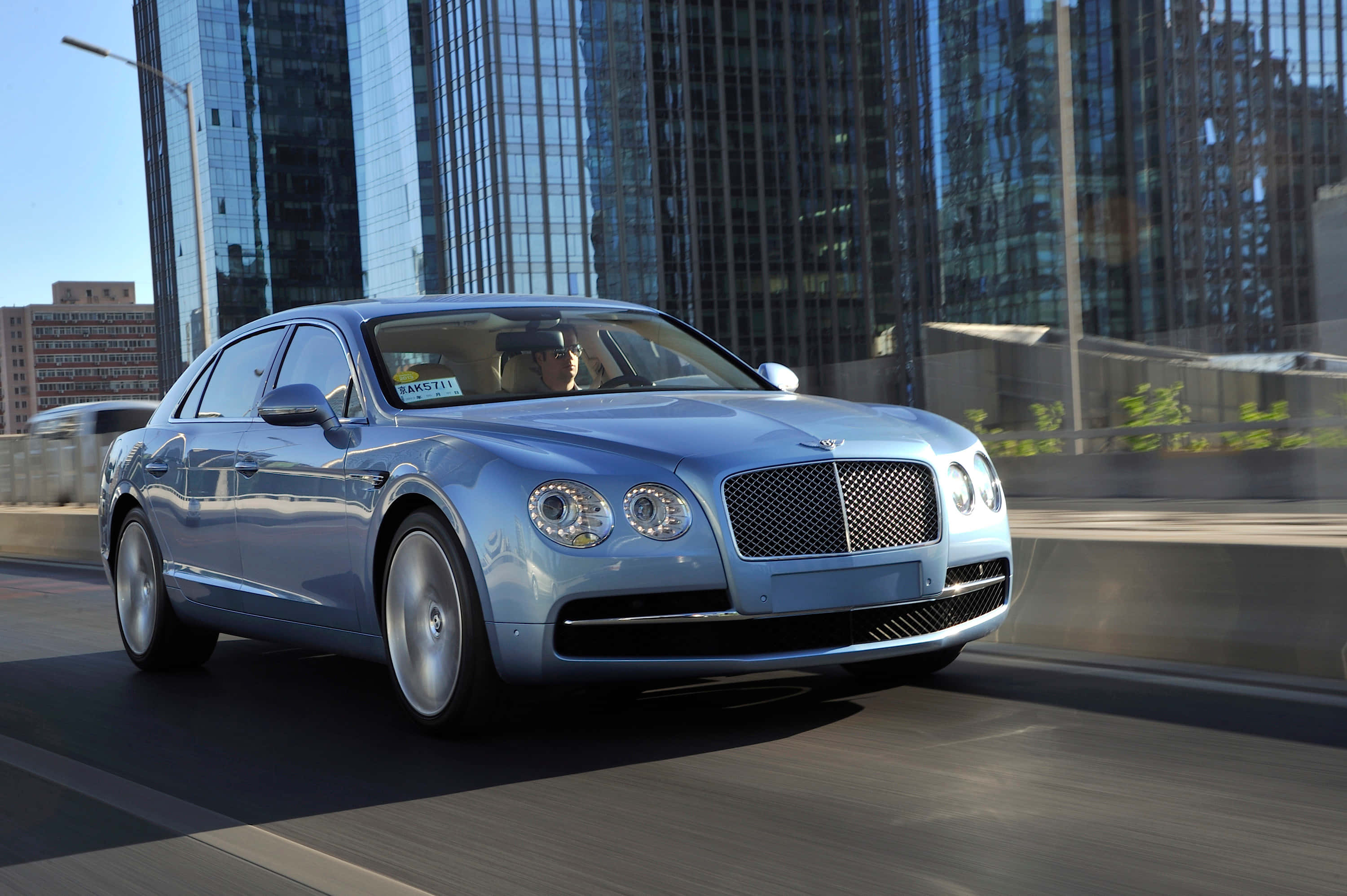 Bentley Flying Spur 桌布 壁紙 — 一輛時尚的銀色賓利轎車在城市高速公路上行駛，高聳的玻璃摩天大樓在其拋光表面上反射。