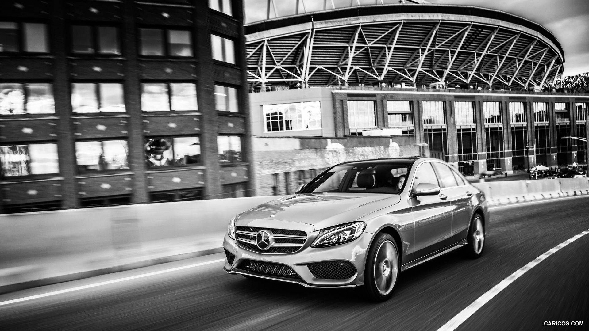 Mercedes Benz C300 桌布 壁紙 — 一輛時尚的銀色賓士轎車在動感的黑白城市鏡頭中，飛馳經過現代體育館和玻璃建築。
