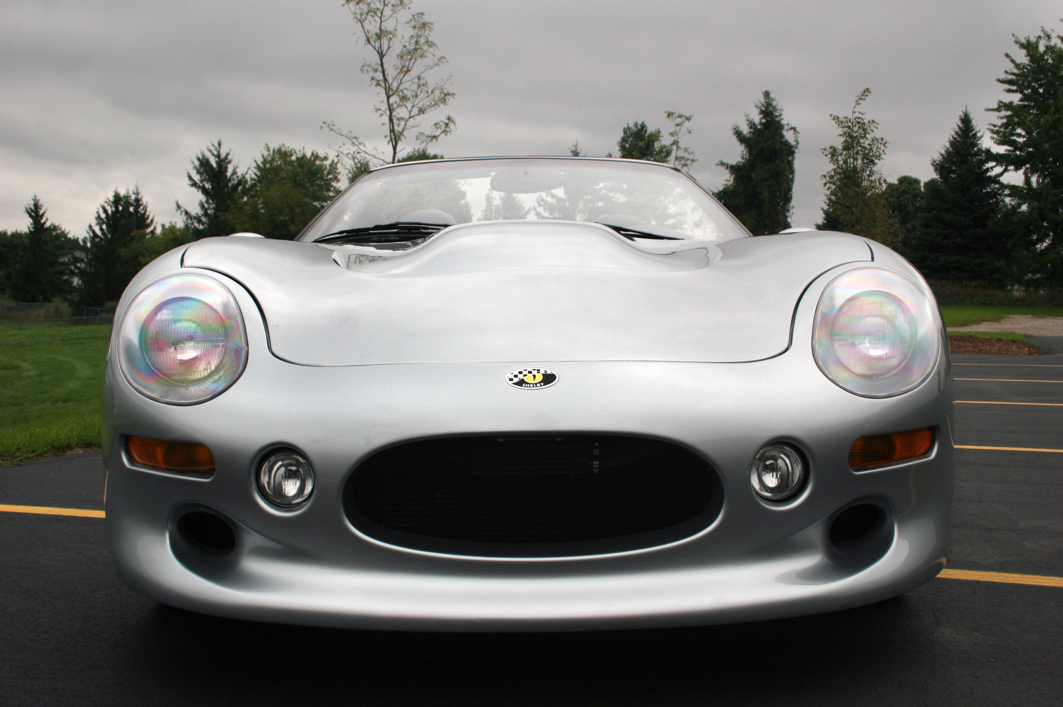 shelby iPhone 桌布 壁紙 — 一輛時尚的銀色跑車，帶有彩虹色車頭燈，停在陰天天空下的瀝青路面上。