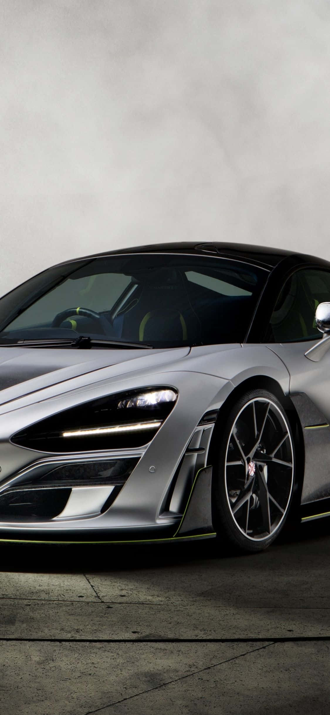iPhone Xs Mclaren 720s 桌布 壁紙 — 一輛時尚的銀色跑車，帶有發光頭燈和黑色飾邊，停在混凝土表面上，背景是柔和的灰色牆壁。
