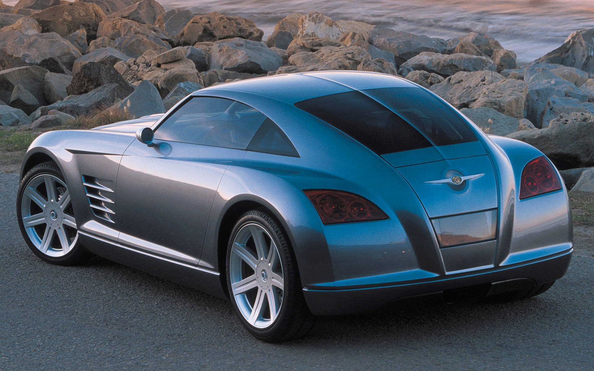 Chrysler 桌布 壁紙 — 一輛時尚的銀色跑車，帶有紅色尾燈，在日落時停在水邊大岩石旁的柏油路面上。