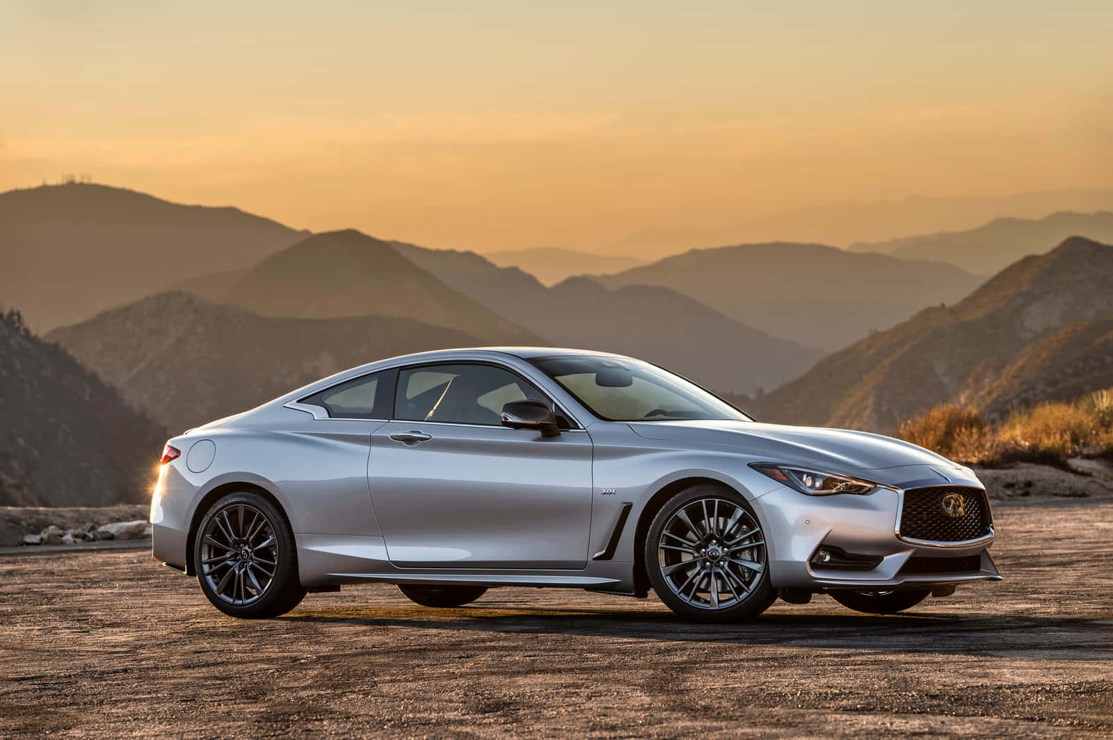 Infiniti Q60 桌布 壁紙 — 一輛時尚的銀色跑車停在山路上，正值黃金時刻，背景是層層疊疊的山丘。