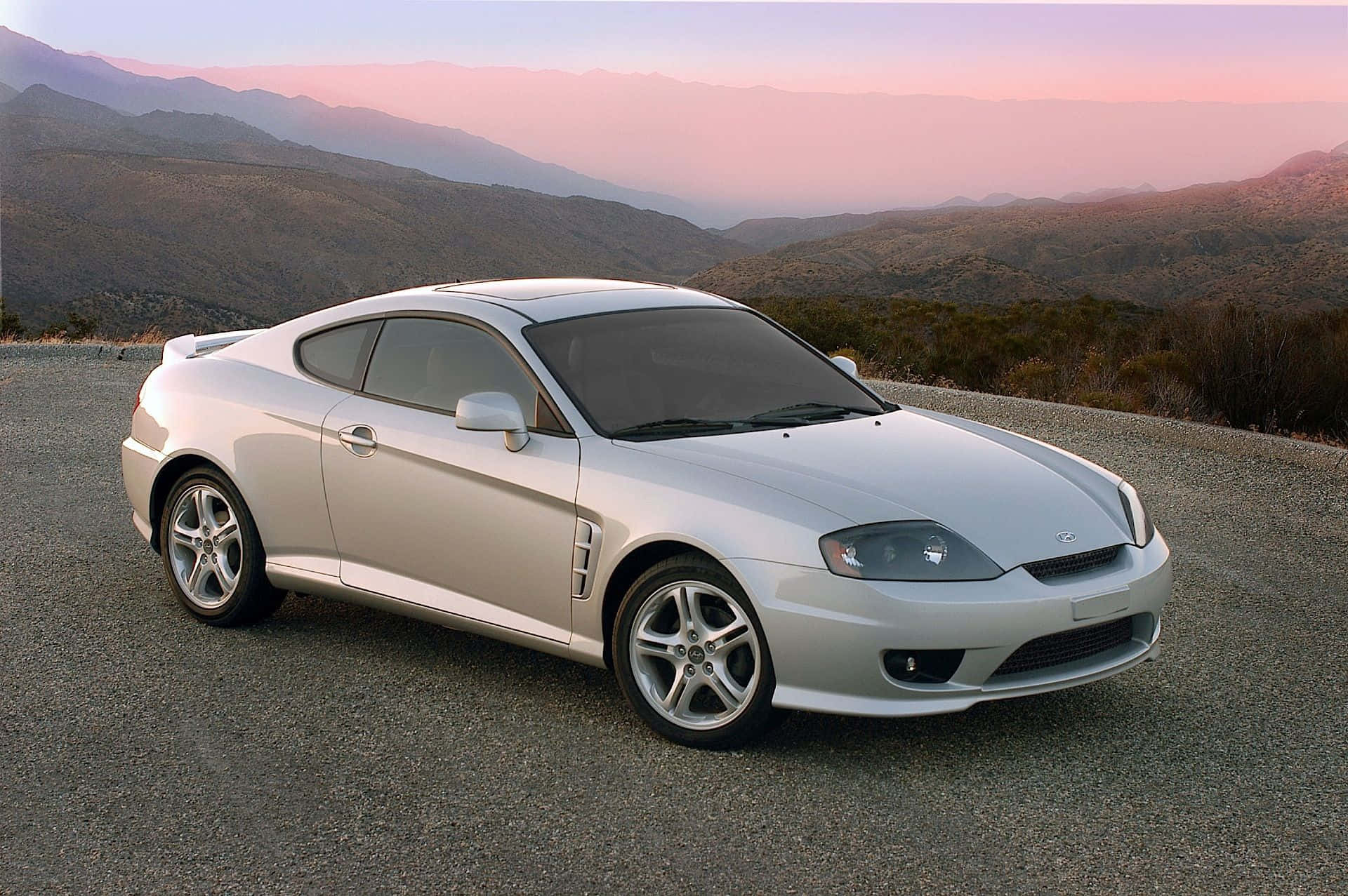 Hyundai Tiburon 桌布 壁紙 — 一輛時尚的銀色跑車停在蜿蜒的山路上，夕陽映照著粉紅和橙色的天空。