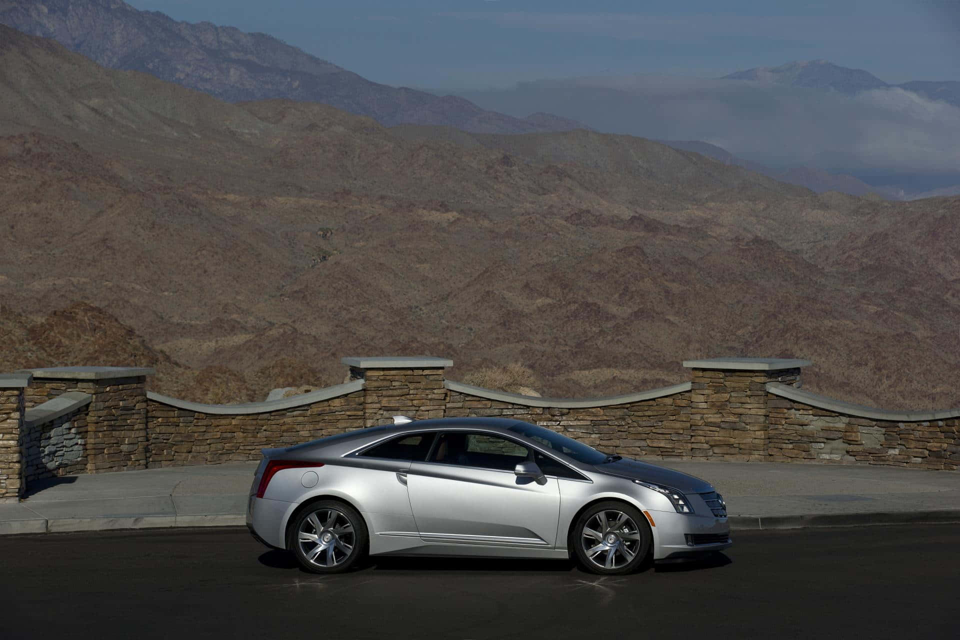 Cadillac Elr 桌布 壁紙 — 一輛時尚的銀色跑車停放在路邊觀景點，背景是崎嶇的棕色山脈和石牆，天空晴朗。