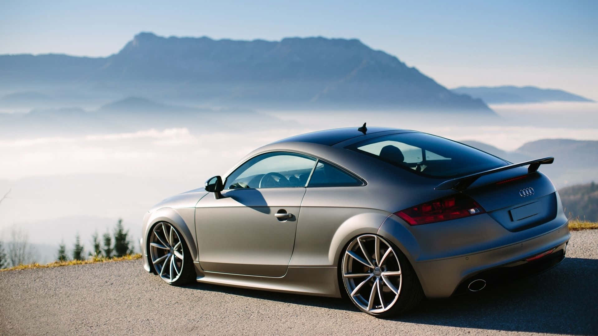 Audi Tt 桌布 壁紙 — 一輛時尚的銀色跑車停泊在山路上，俯瞰著一片霧海和遠處的山峰，天空清澈湛藍。