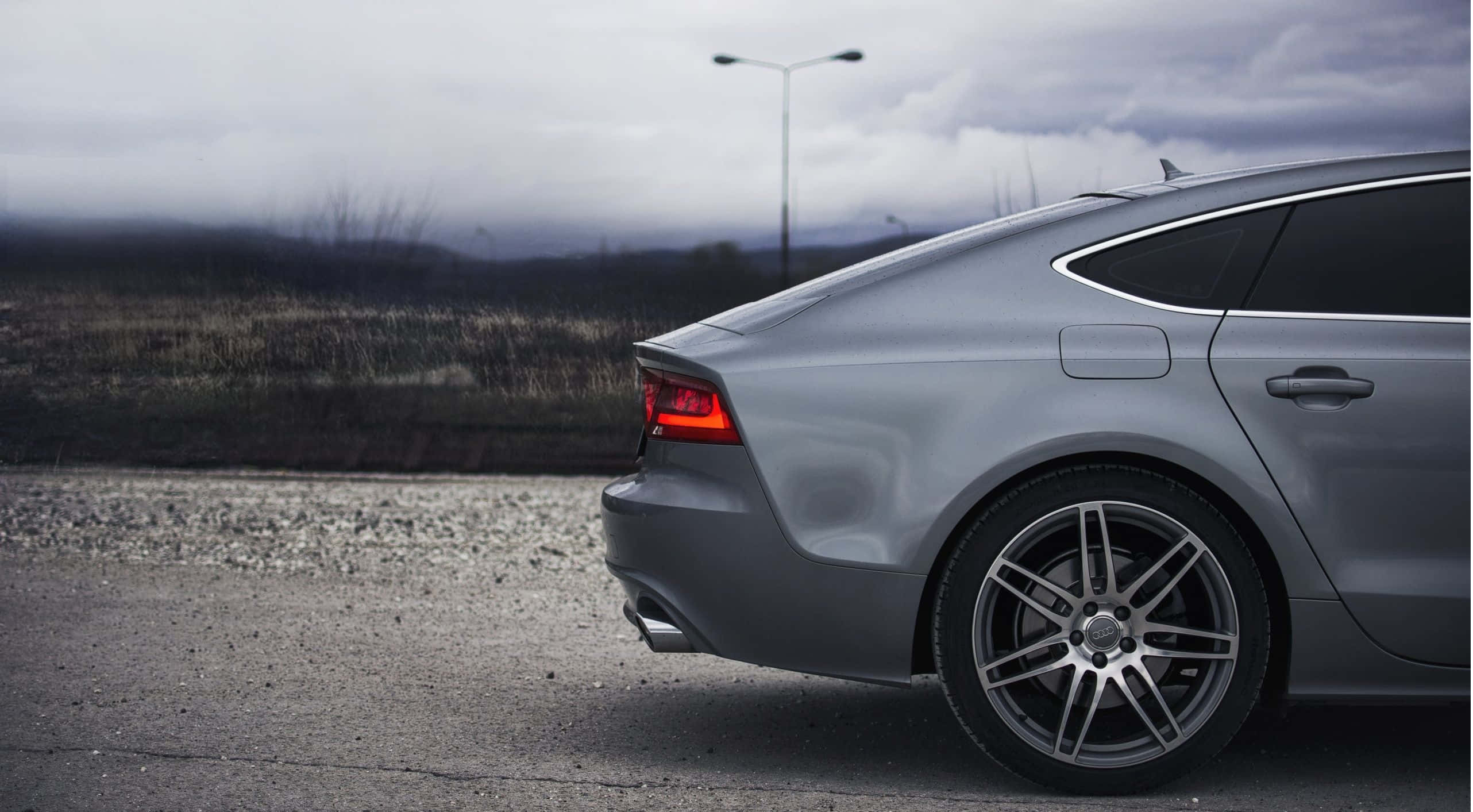 Audi S7 桌布 壁紙 — 一輛時尚的銀色跑車停泊在碎石路邊，天空多雲，遠處有山丘。