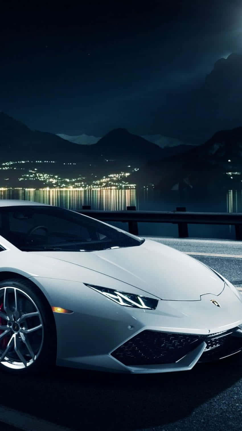 Pixel 3xl Lamborghini 桌布 壁紙 — 一輛時尚的銀色跑車在夜燈下閃耀，旁邊是反射的湖泊，遠處的山城在背景中閃爍。