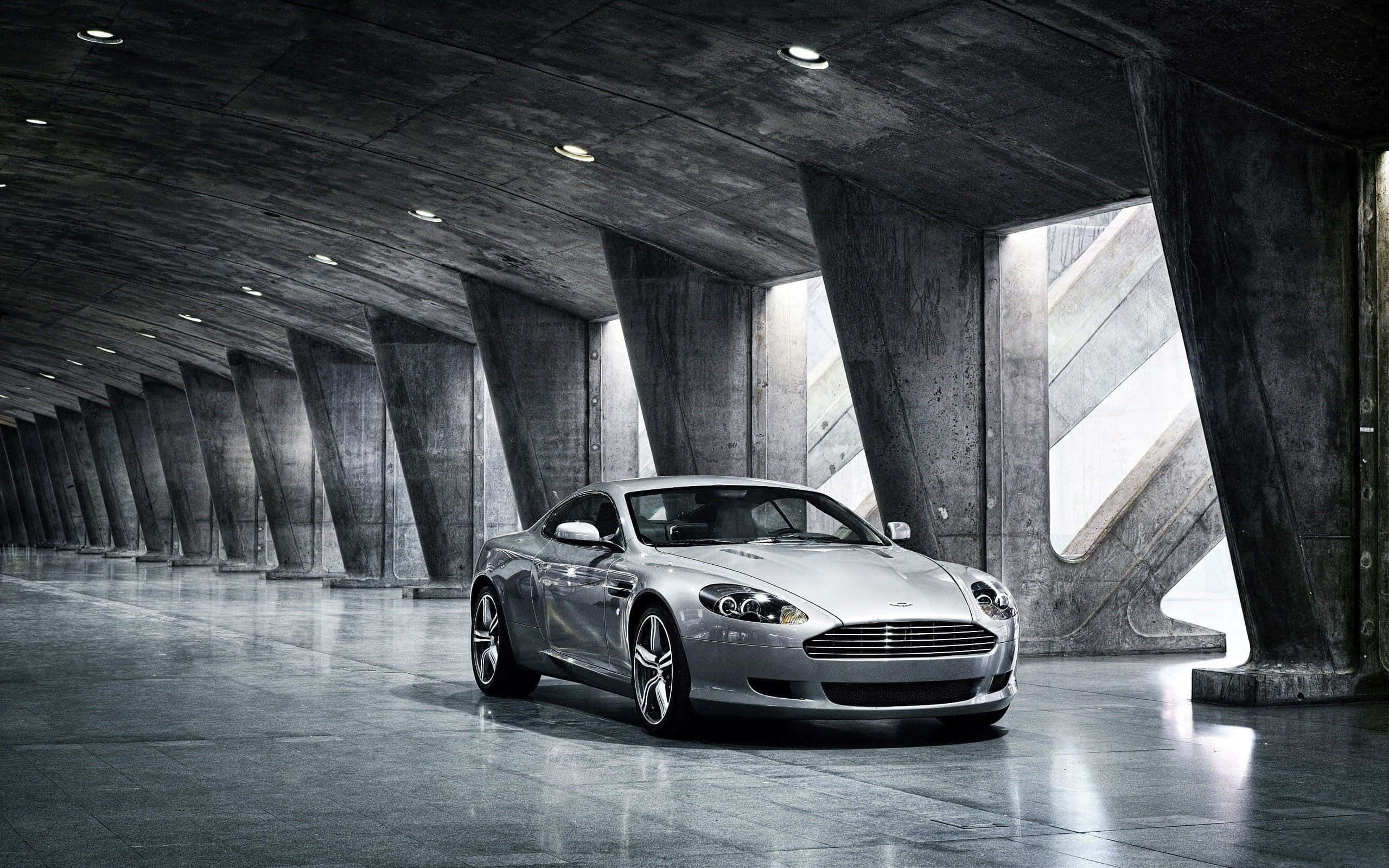 Aston Martin Db9 桌布 壁紙 — 一輛時尚的銀色跑車在寬敞的混凝土拱形車庫中，在工業燈光下閃閃發光。