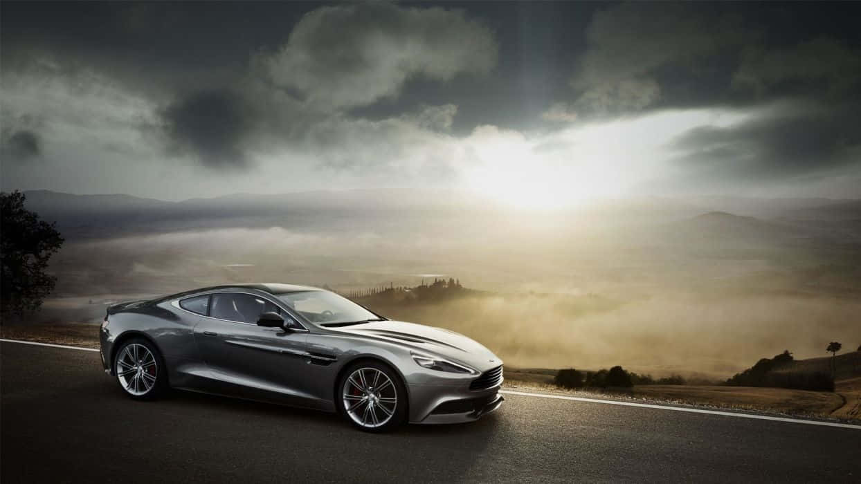 Aston Martin Vanquish 桌布 壁紙 — 一輛時尚的銀色跑車在戲劇性的多雲天空下，沿著蜿蜒的山路行駛，陽光穿透雲層。