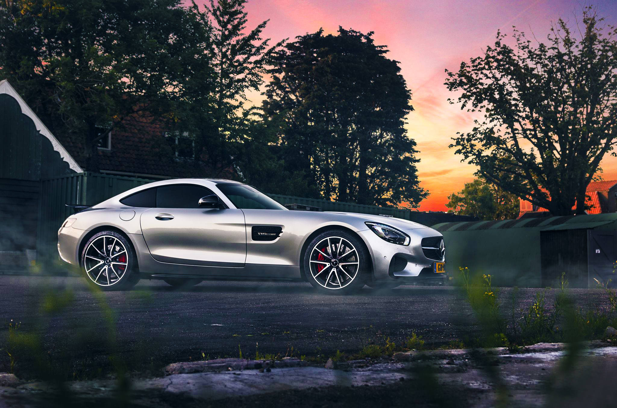Mercedes Benz 桌布 壁紙 — 一輛時尚的銀色跑車在日落時分穿過薄霧，身後是樹葉茂密的樹木，天空呈現火熱的橙色和粉紅色。