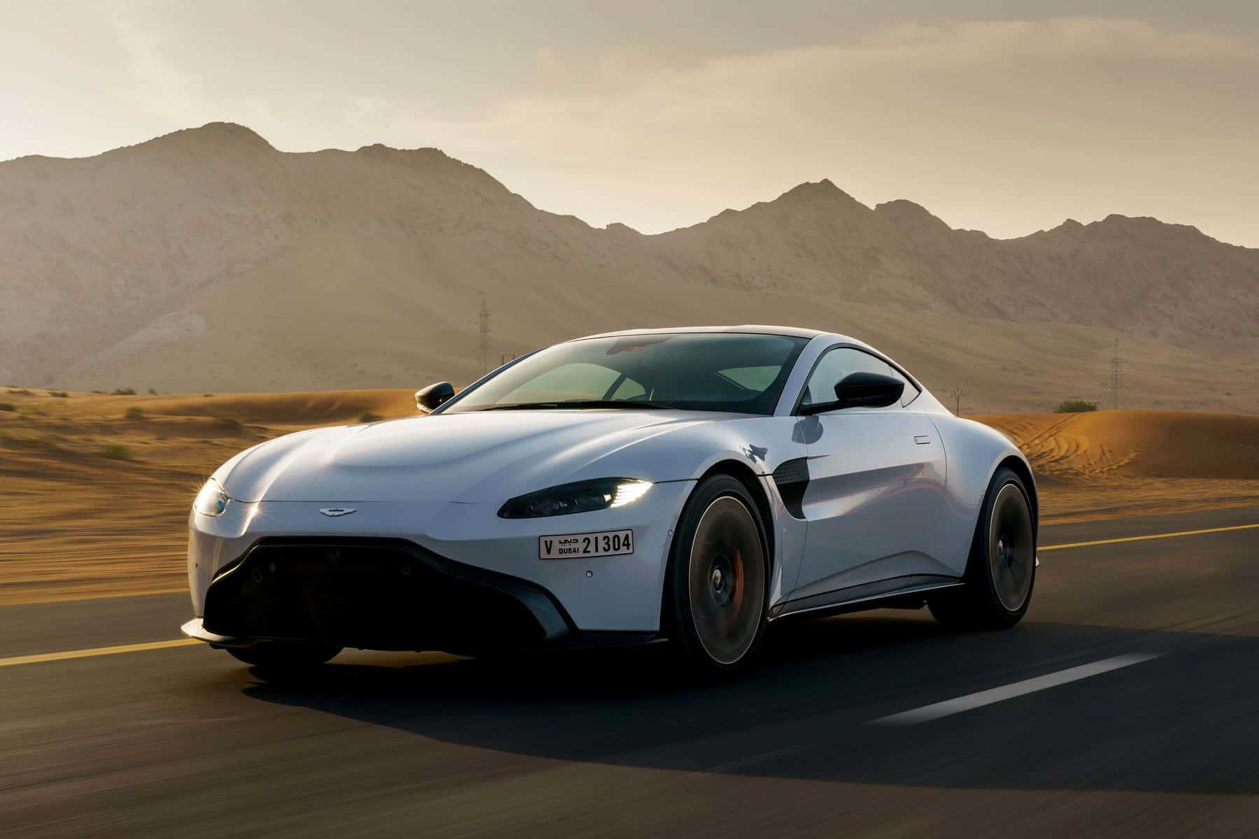 Aston Martin Vantage 桌布 壁紙 — 一輛時尚的銀色跑車在沙漠公路上疾馳，沿途有沙丘和遠山，天空溫暖金黃。