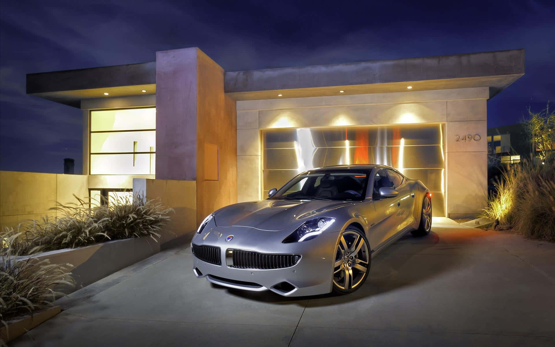 Fisker 桌布 壁紙 — 一輛時尚的銀色跑車在溫暖的車庫燈光下閃閃發光，停放在黃昏時分的現代混凝土房屋前。