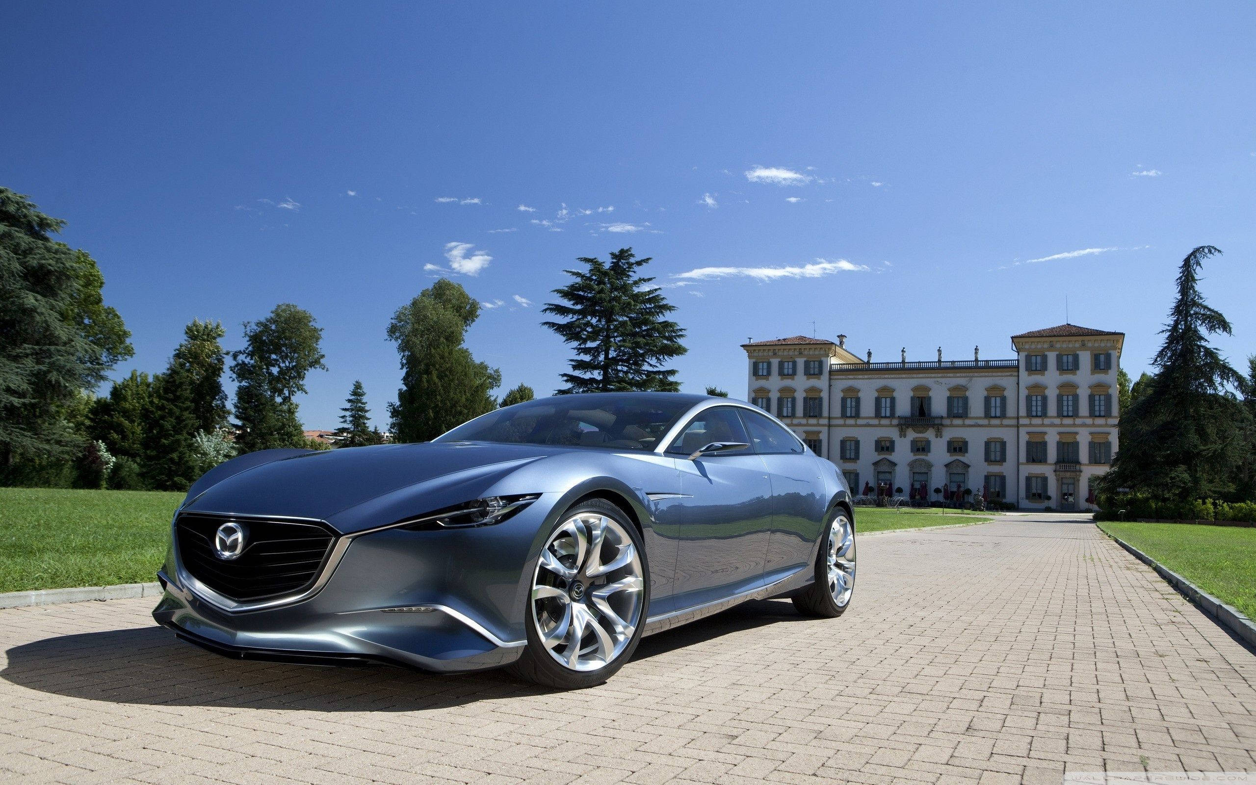 Mazda 桌布 壁紙 — 一輛時尚的銀色跑車在燦爛陽光下閃閃發光，停在石頭車道上，前面是一座宏偉的歐洲別墅，周圍環繞著茂密的綠樹。