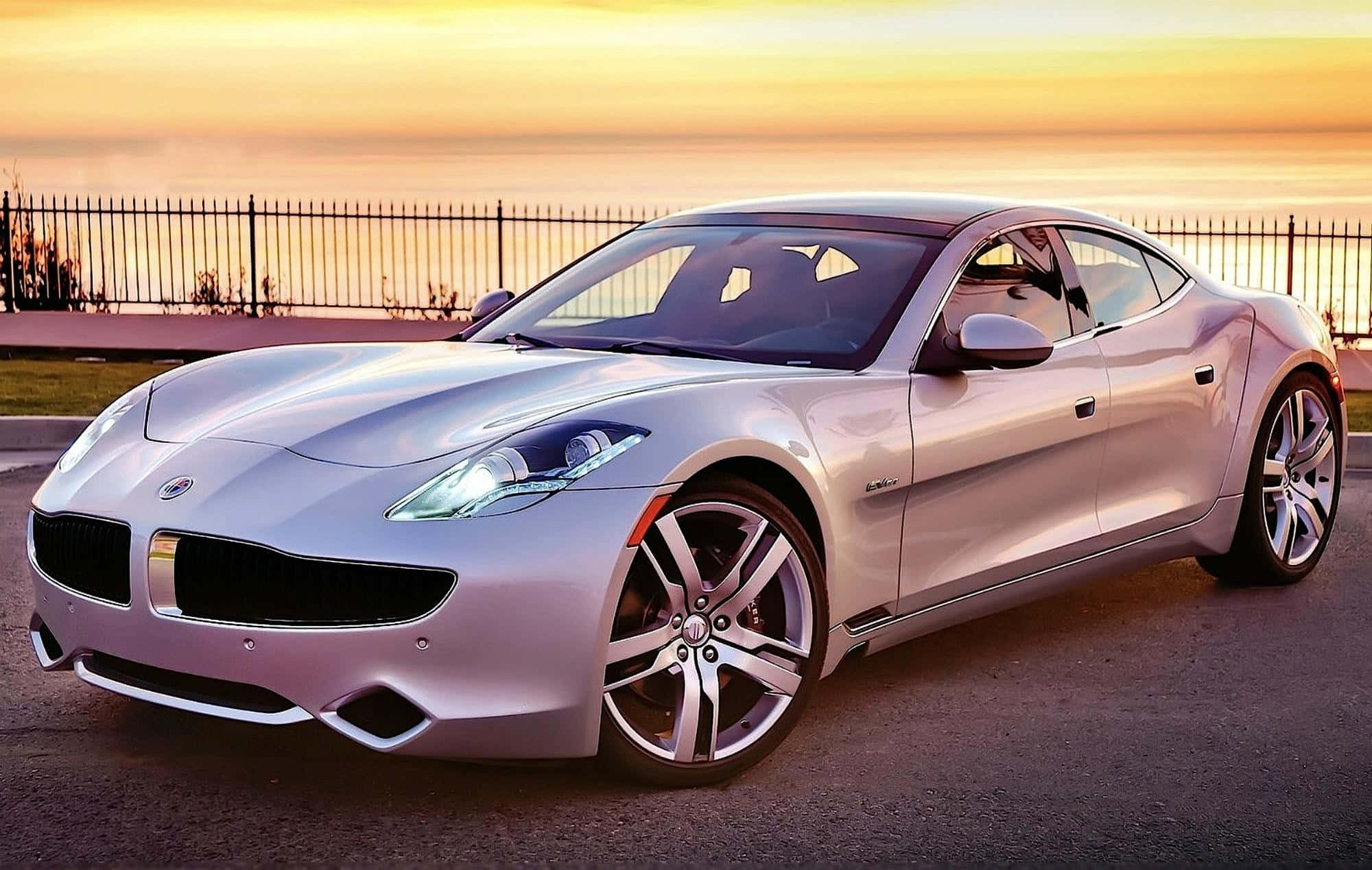 Fisker 桌布 壁紙 — 一輛時尚的銀色跑車在金色夕陽下閃閃發光，停泊在俯瞰水面的圍欄旁。