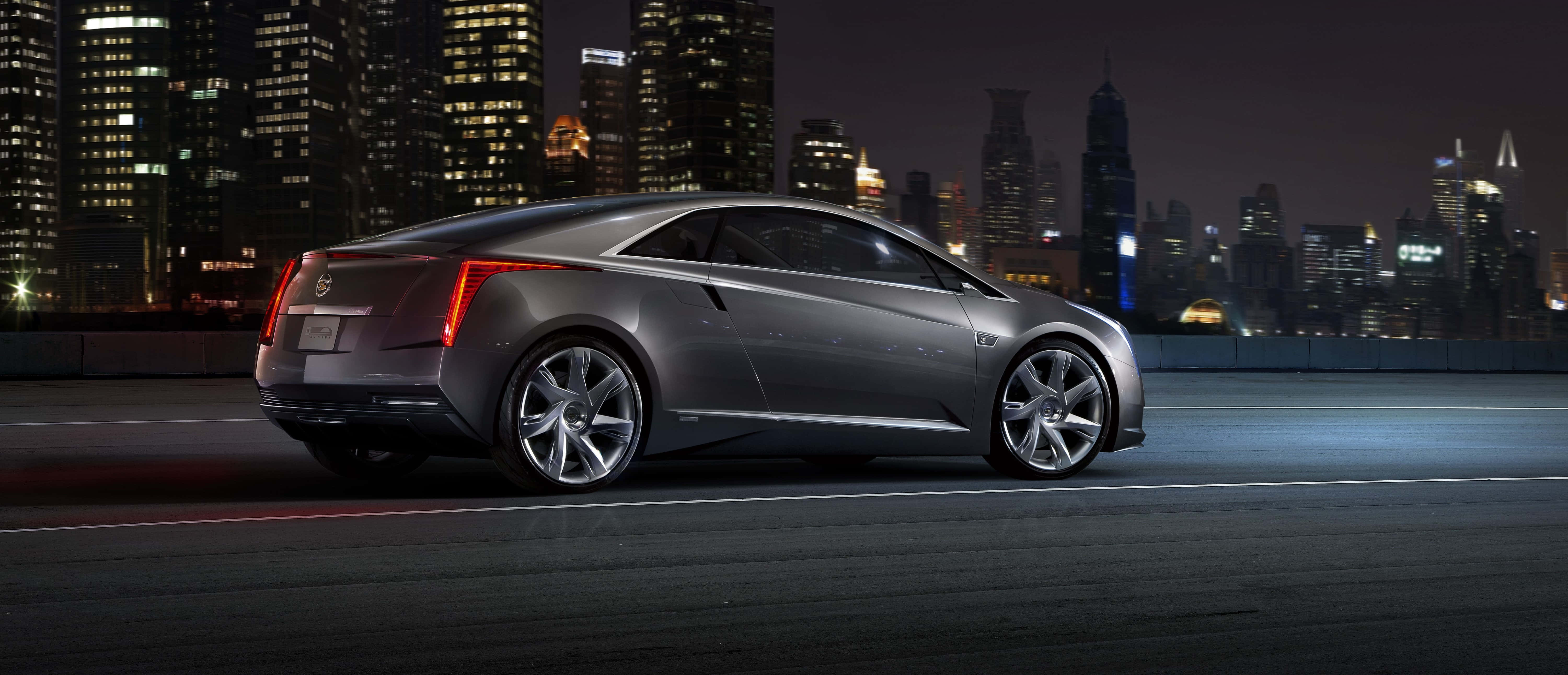 Cadillac Elr 桌布 壁紙 — 一輛時尚的銀色跑車在閃爍的夜空下，沿著城市高速公路行駛，背後是高聳的摩天大樓。