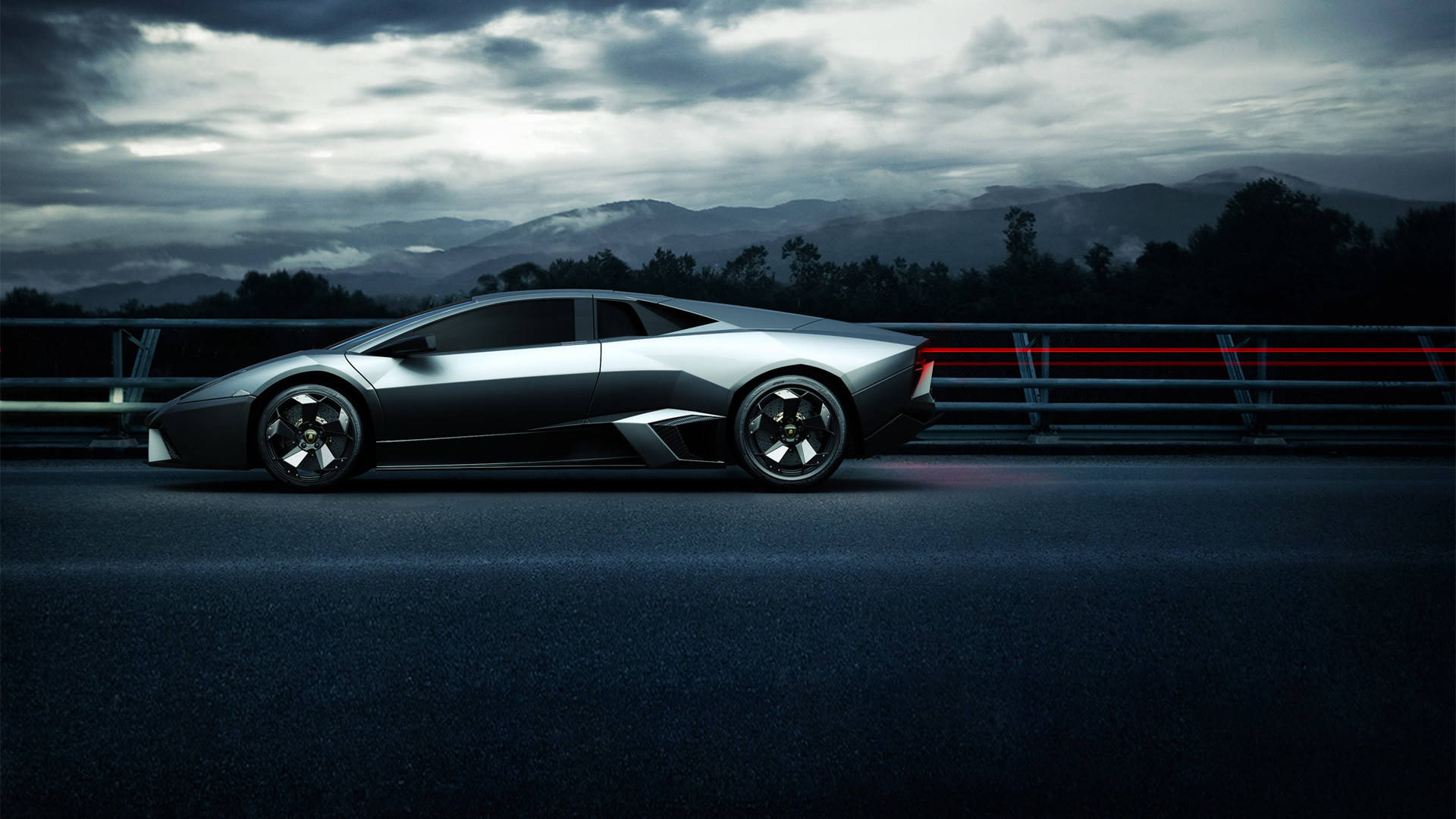 Lamborghini 4K 桌布 壁紙 — 一輛時尚的銀色跑車在陰暗多雲的天空下沿著黑暗的高速公路行駛，遠處有霧氣繚繞的山巒。