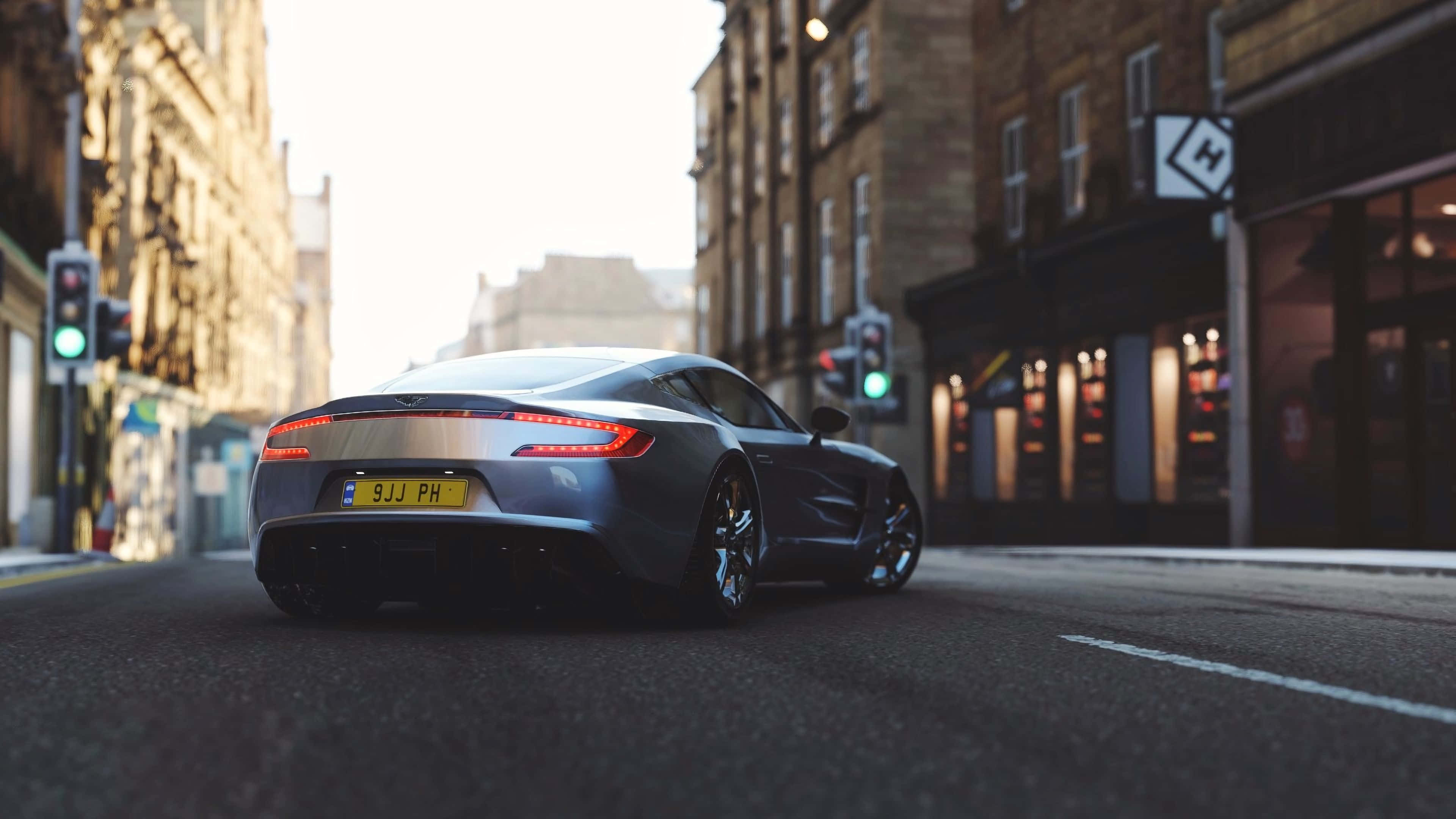 Aston Martin One-77 桌布 壁紙 — 一輛時尚的銀色跑車在陽光普照的市區街道上滑行，兩旁是歷史悠久的磚造建築和閃閃發光的商店櫥窗。