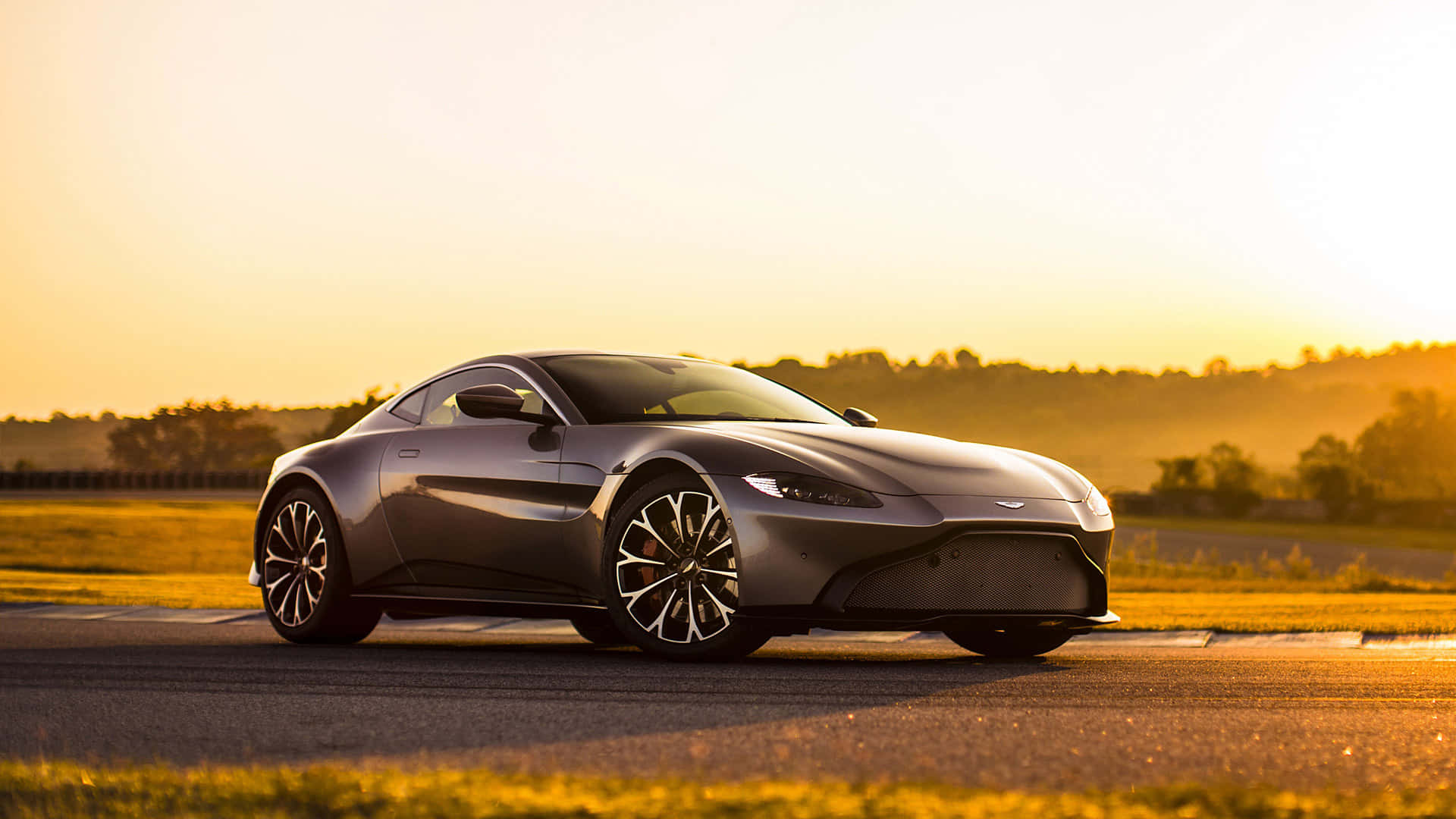 Aston Martin Vantage 桌布 壁紙 — 一輛時尚的銀色跑車在陽光普照的賽道上滑行，金色光芒以溫暖色調沐浴著風景。