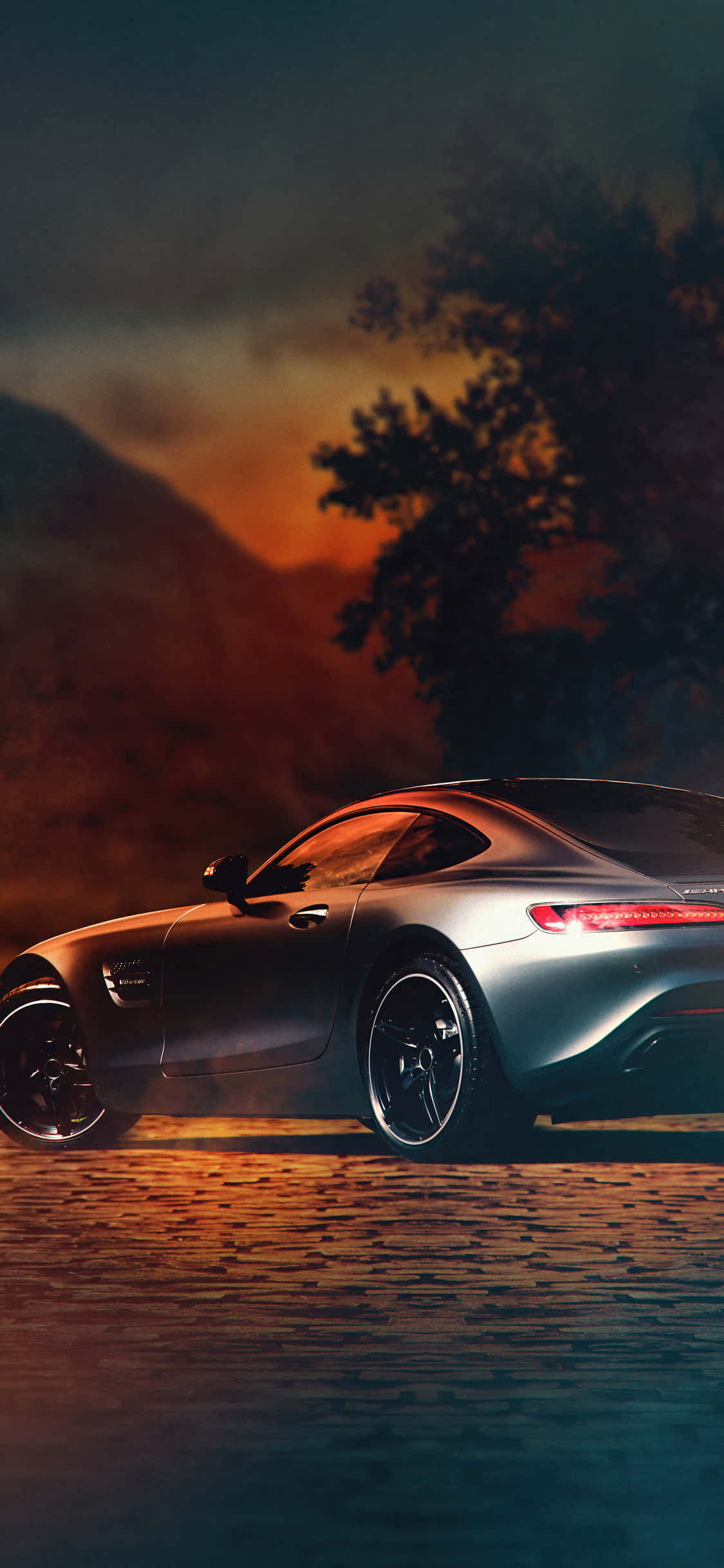 iPhone X Amg Gt-r 桌布 壁紙 — 一輛時尚的銀色跑車在鵝卵石路上行駛，天空是火紅的日落，襯托出樹木和山脈的剪影。