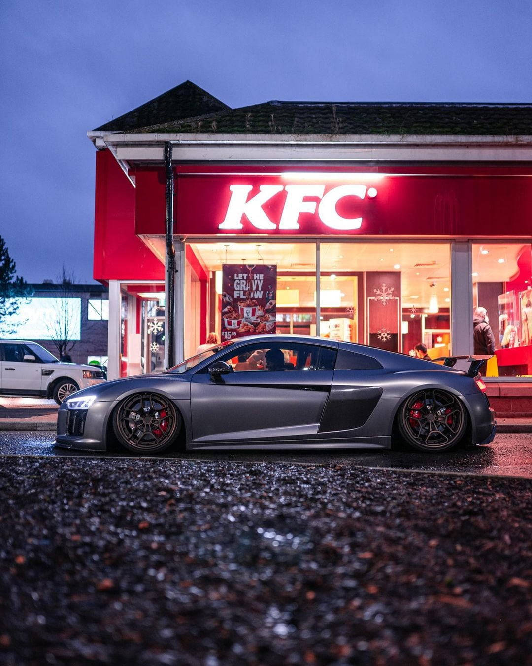 肯德基 桌布 壁紙 — 一輛時尚的銀色跑車在黃昏時停在一間燈火通明的KFC餐廳前，潮濕的路面反射著霓虹燈光。