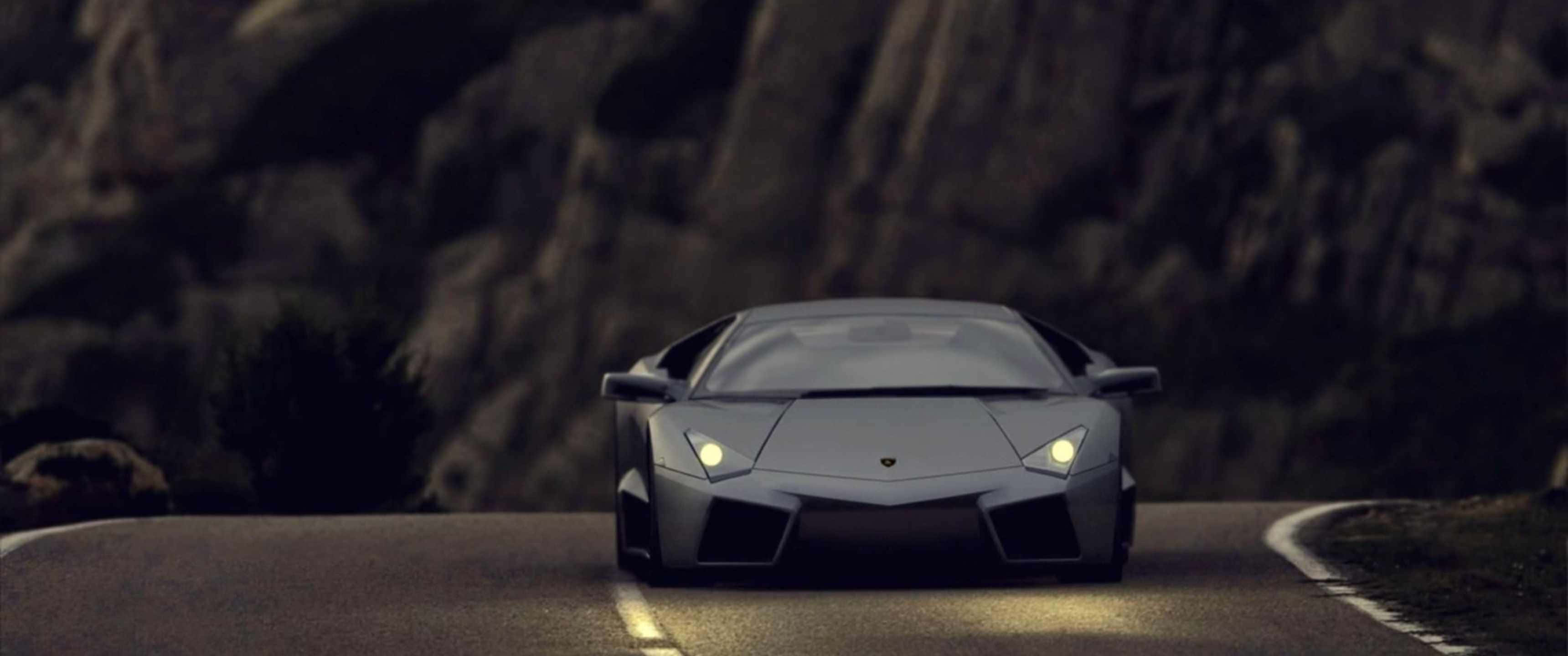 3440x1440p Lamborghini 桌布 壁紙 — 一輛時尚的銀色跑車在黃昏時分沿著蜿蜒的山路行駛，車頭燈閃閃發光。
