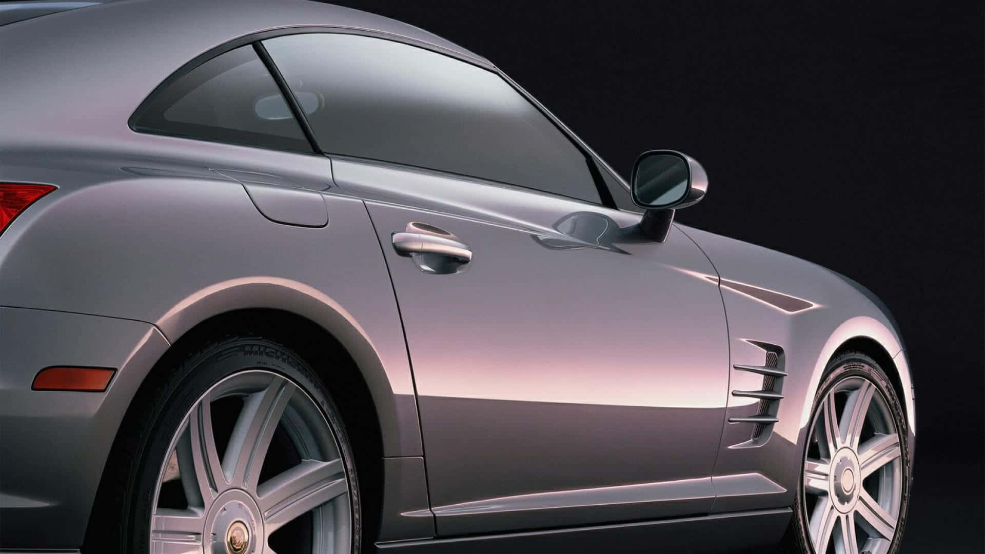 Chrysler Crossfire 桌布 壁紙 — 一輛時尚的銀色跑車在黑暗的攝影棚中滑行，擁有拋光曲線和閃亮的合金輪圈。