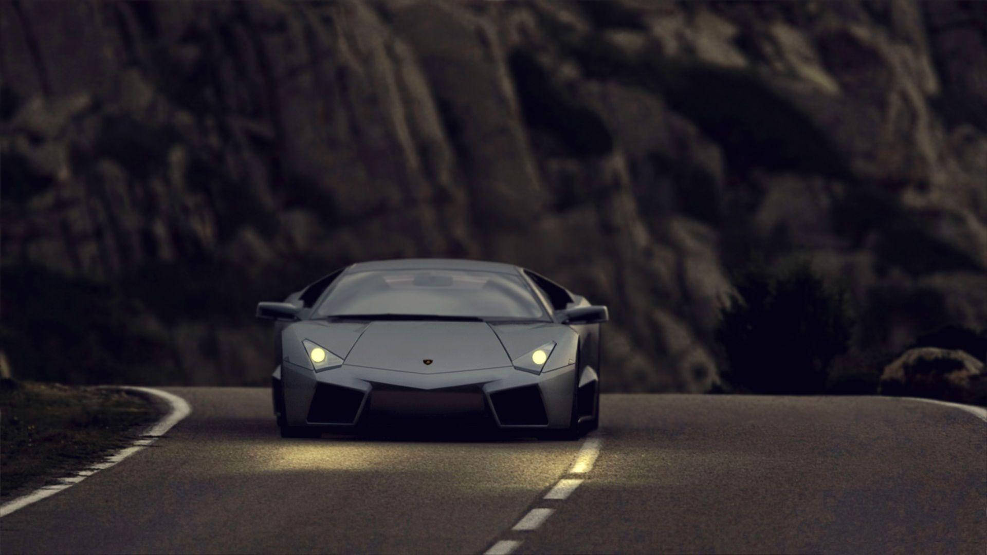 Lamborghini 4K 桌布 壁紙 — 一輛時尚的銀色跑車帶著發光的車頭燈，在黃昏時分沿著蜿蜒的山路駛向觀眾。