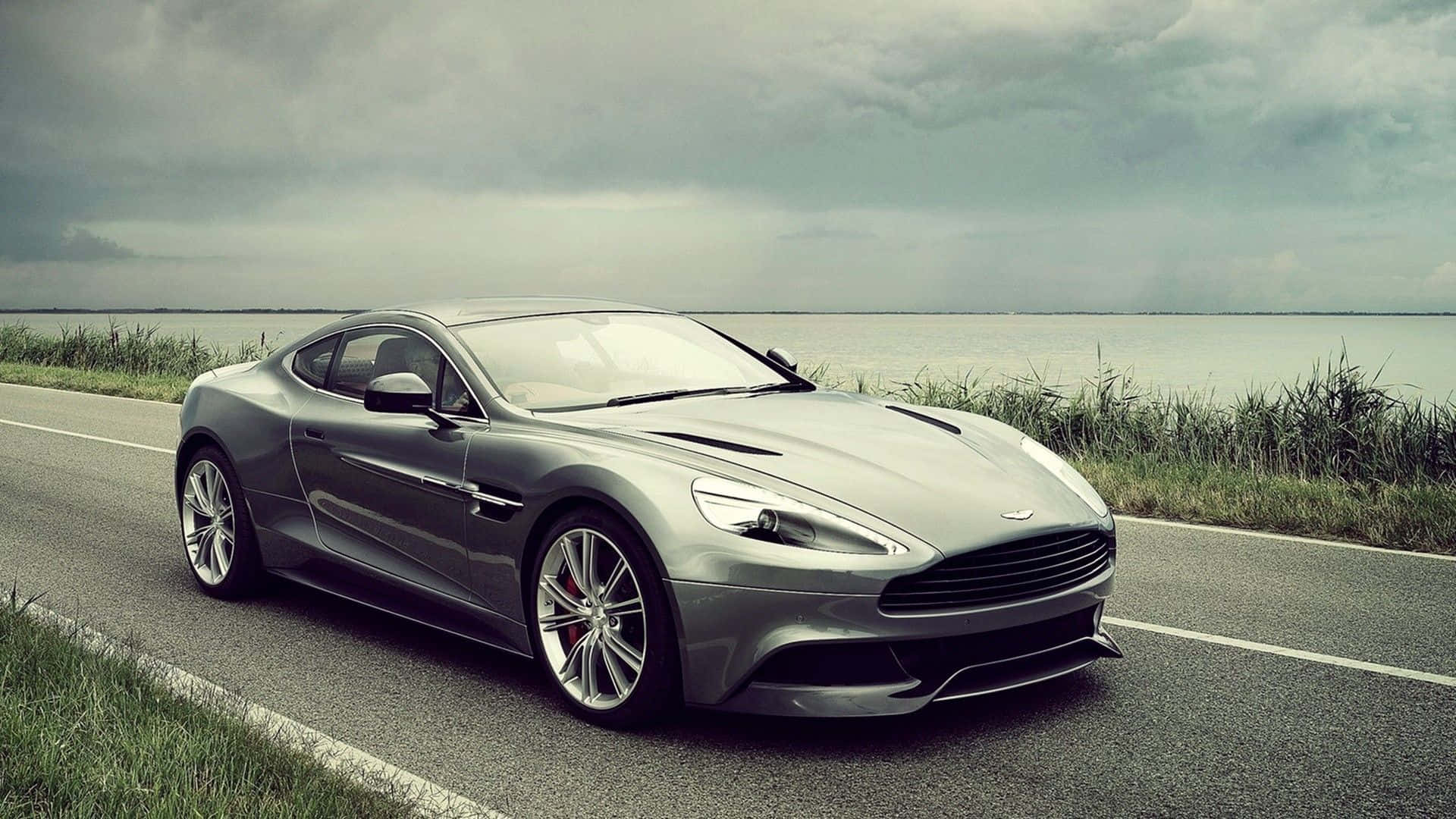 Aston Martin Vanquish 桌布 壁紙 — 一輛時尚的銀色跑車沿著海岸公路行駛，背景是陰沉的天空、蘆葦和水面。