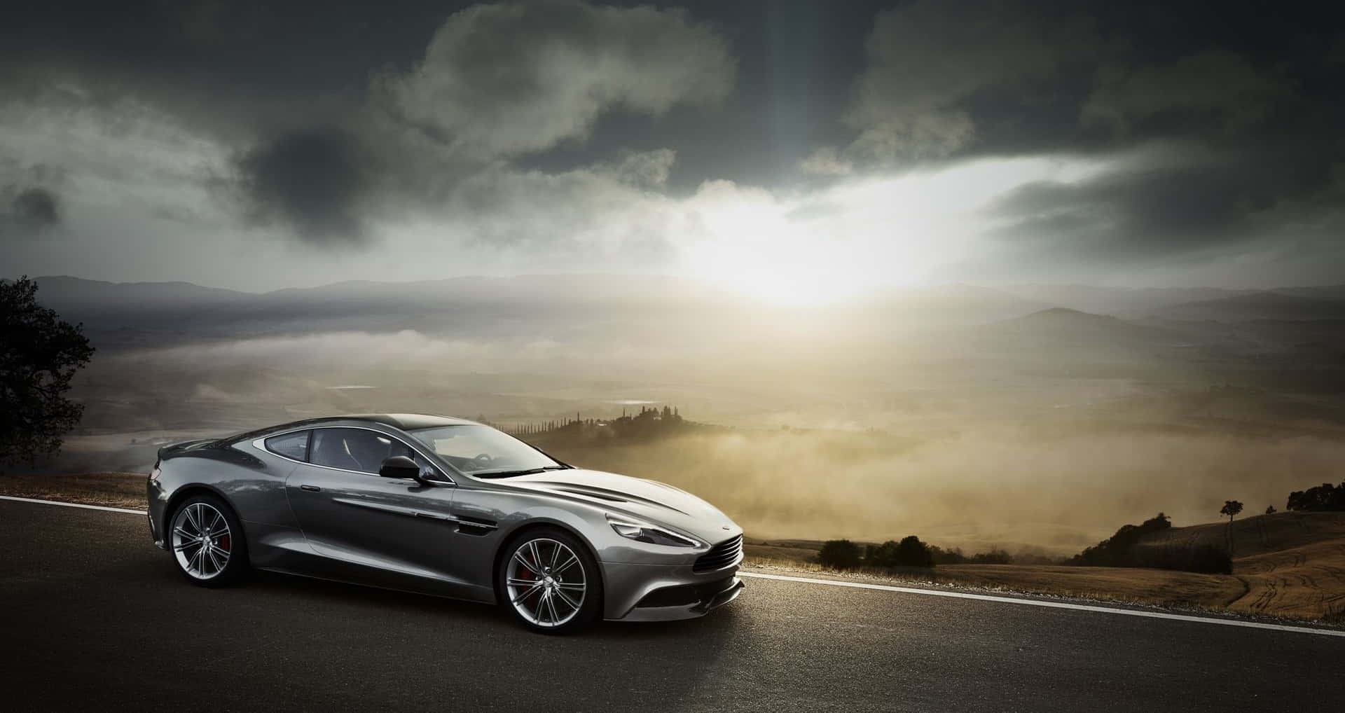 Aston Martin Db9 桌布 壁紙 — 一輛時尚的銀色跑車沿著蜿蜒的道路行駛，穿過霧氣籠罩的起伏山丘，天空戲劇性地佈滿雲層。