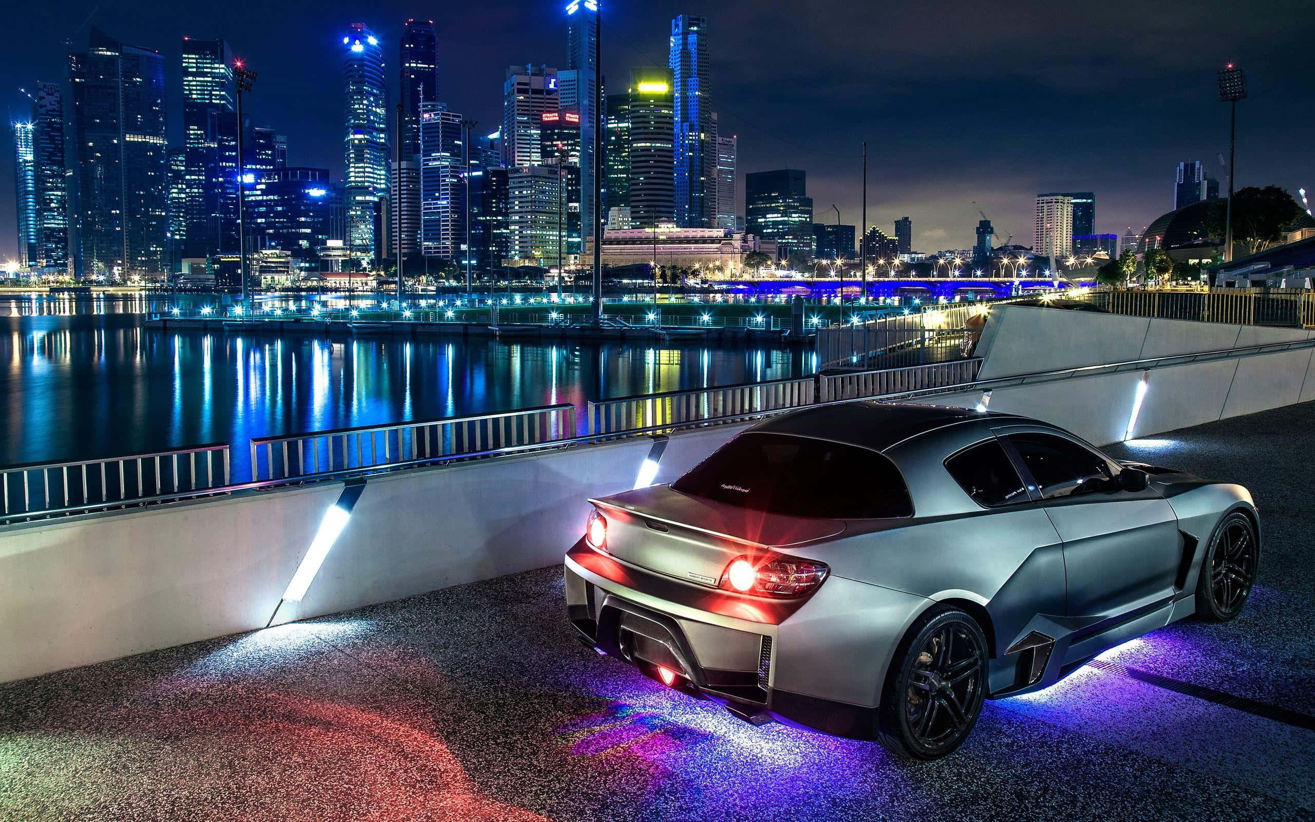 Mazda Rx-8 桌布 壁紙 — 一輛時尚的銀色跑車發出紫色底盤燈光，停在夜間的海濱長廊上，映照著充滿活力的城市天際線。