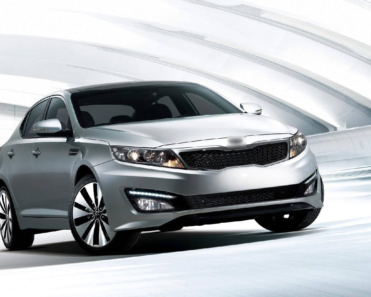 Kia 桌布 壁紙 — 一輛時尚的銀色轎車，配備銳利的頭燈和時尚的合金輪圈，在未來主義風格的白色彎曲隧道背景下蓄勢待發。