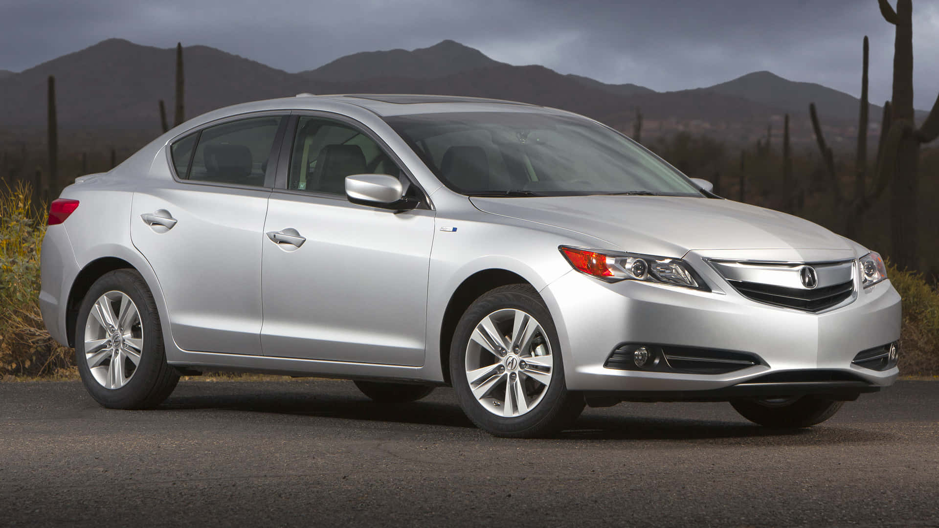 Acura Ilx 桌布 壁紙 — 一輛時尚的銀色轎車停在柏油路上，背景是山脈和仙人掌，天空多雲。